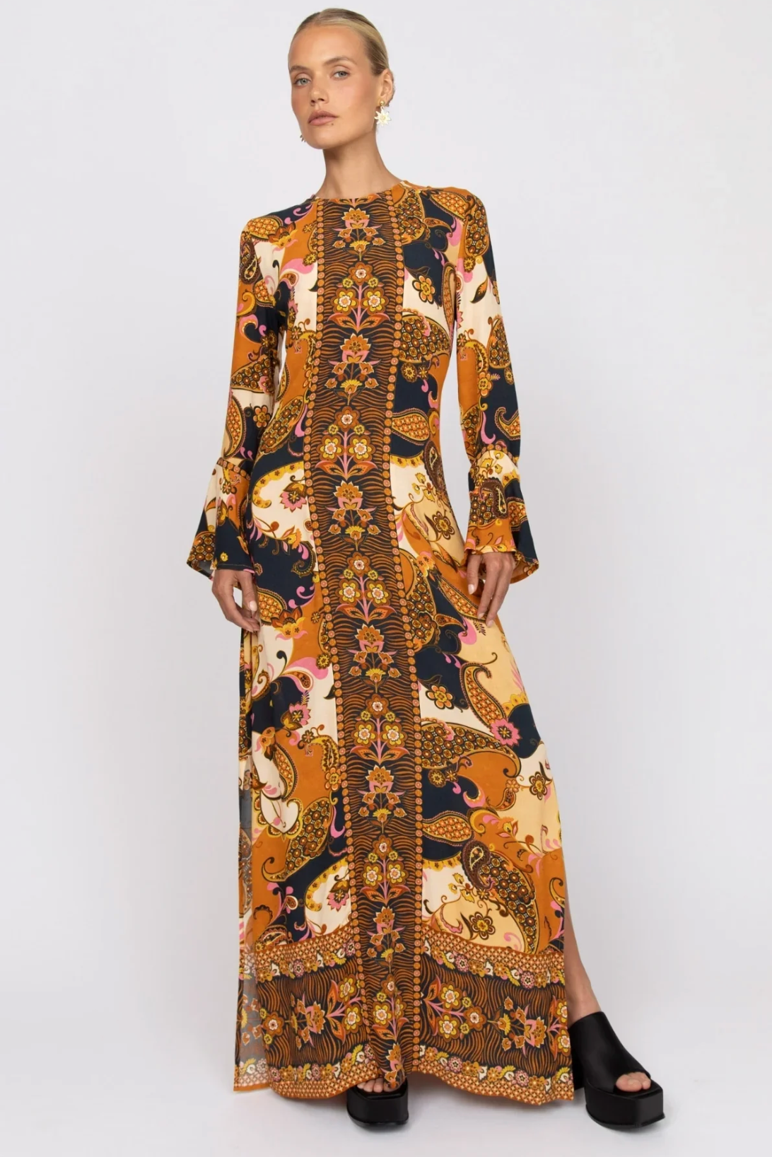 A woman in the Nine Lives Bazaar Citrine Gown - Cocoa, a long special occasion dress with fluted sleeves and vibrant orange, yellow, black, and brown patterns, stands against a plain white background.