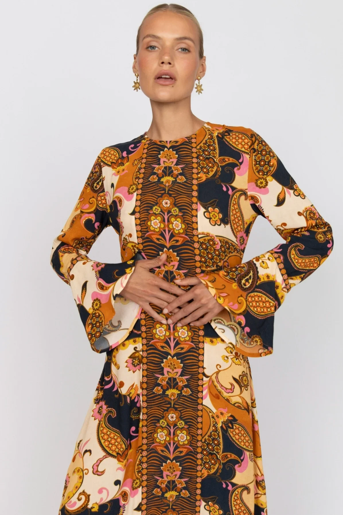 A woman in the Nine Lives Bazaar Citrine Gown - Cocoa, a long-sleeve dress with vibrant paisley and floral patterns in orange, yellow, black, and pink, stands against a plain background with hands folded at her waist.