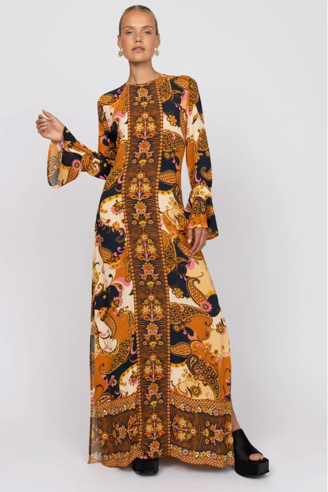 A woman in the Nine Lives Bazaar Citrine Gown - Cocoa, featuring vibrant orange, gold, and black paisley floral patterns and fluted sleeves, pairs it with black platform sandals—an eye-catching special occasion look.