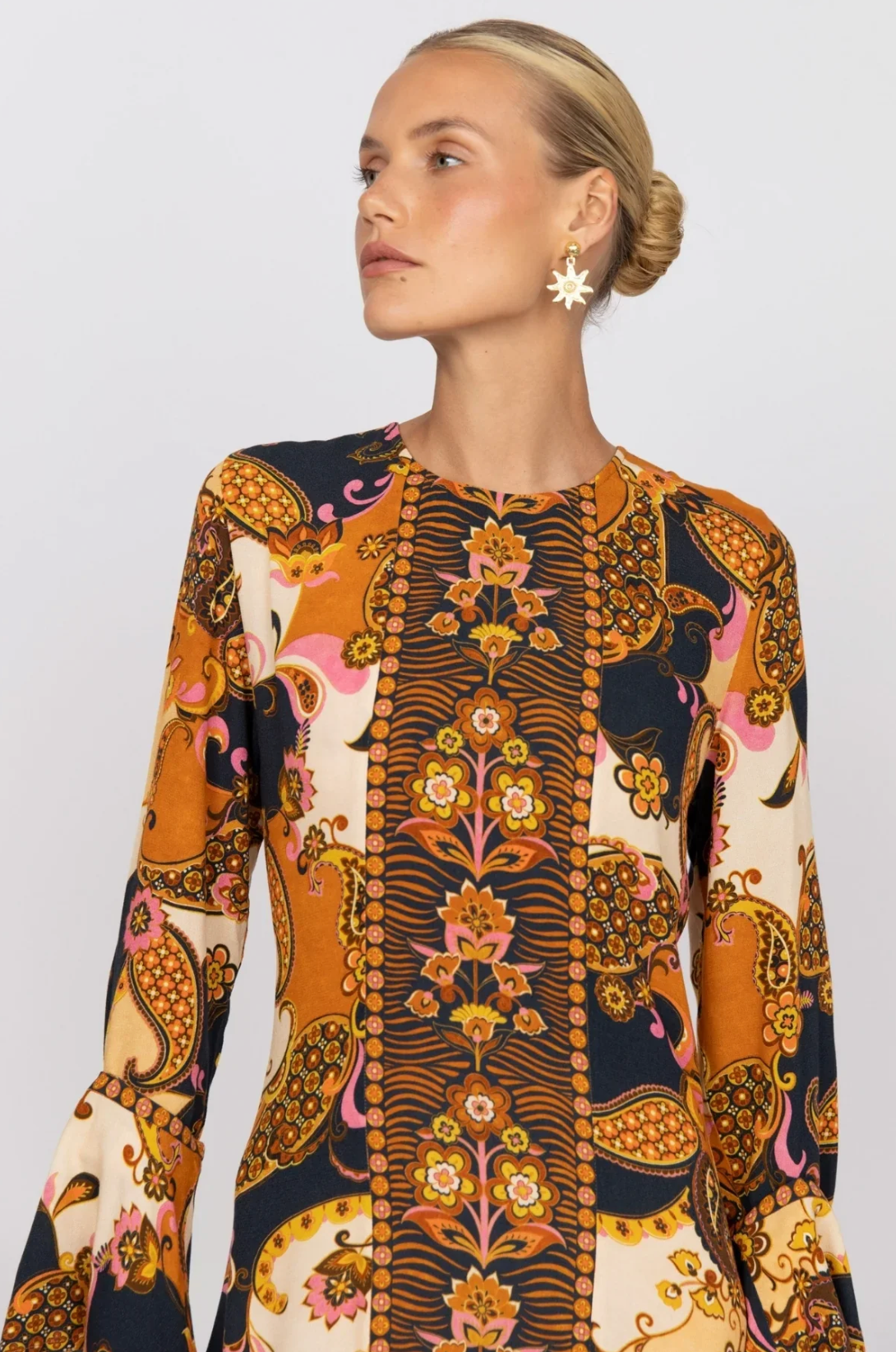 Woman with blonde hair in a low bun, wearing star-shaped earrings and the Nine Lives Bazaar Citrine Gown – Cocoa, a special occasion dress with fluted sleeves and a vibrant paisley and floral print in orange, pink, yellow, navy, and cream tones. Looking to the side.
