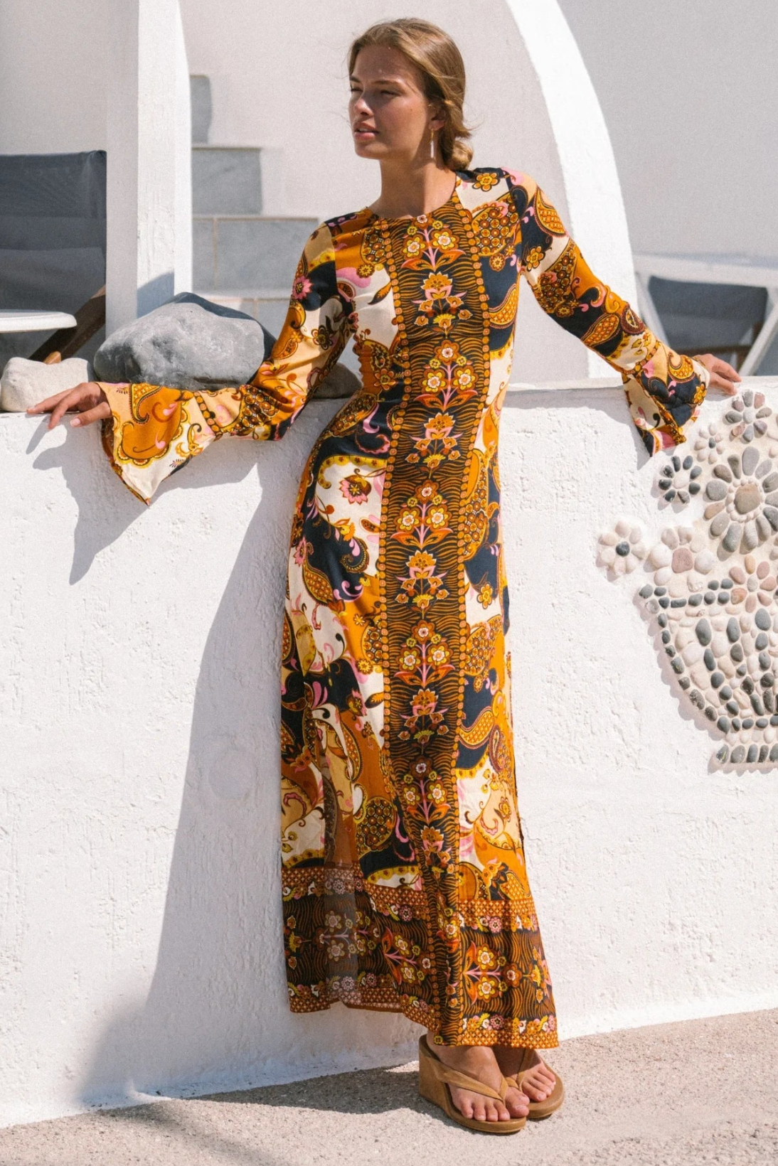 A woman in the Citrine Gown - Cocoa by Nine Lives Bazaar leans against a white outdoor wall, sunlight casting shadows and minimalist white architecture in the background.