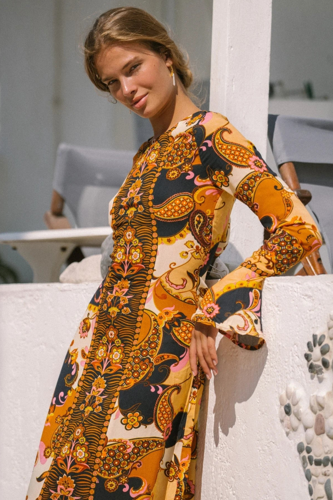 A woman with light brown hair stands outdoors, smiling in the sun as she leans against a white wall, wearing the Citrine Gown - Cocoa by Nine Lives Bazaar with fluted sleeves and vibrant orange, yellow, and black patterns.