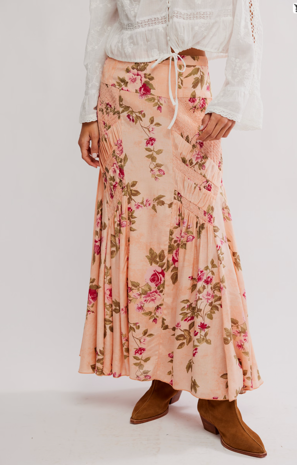 A person wearing a white, long-sleeve blouse and the Rowan Maxi Skirt by Free People in peach with an A-line silhouette and pink rose floral print, paired with brown ankle boots. The image is cropped at the shoulders.