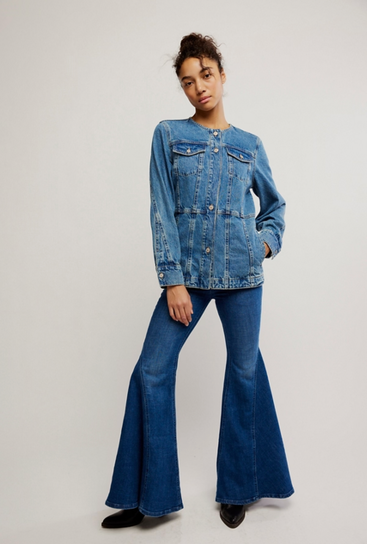 A person stands against a plain background, wearing the Free People Lula Denim Jacket with front pockets and buttons, paired with matching blue flared jeans and black shoes.