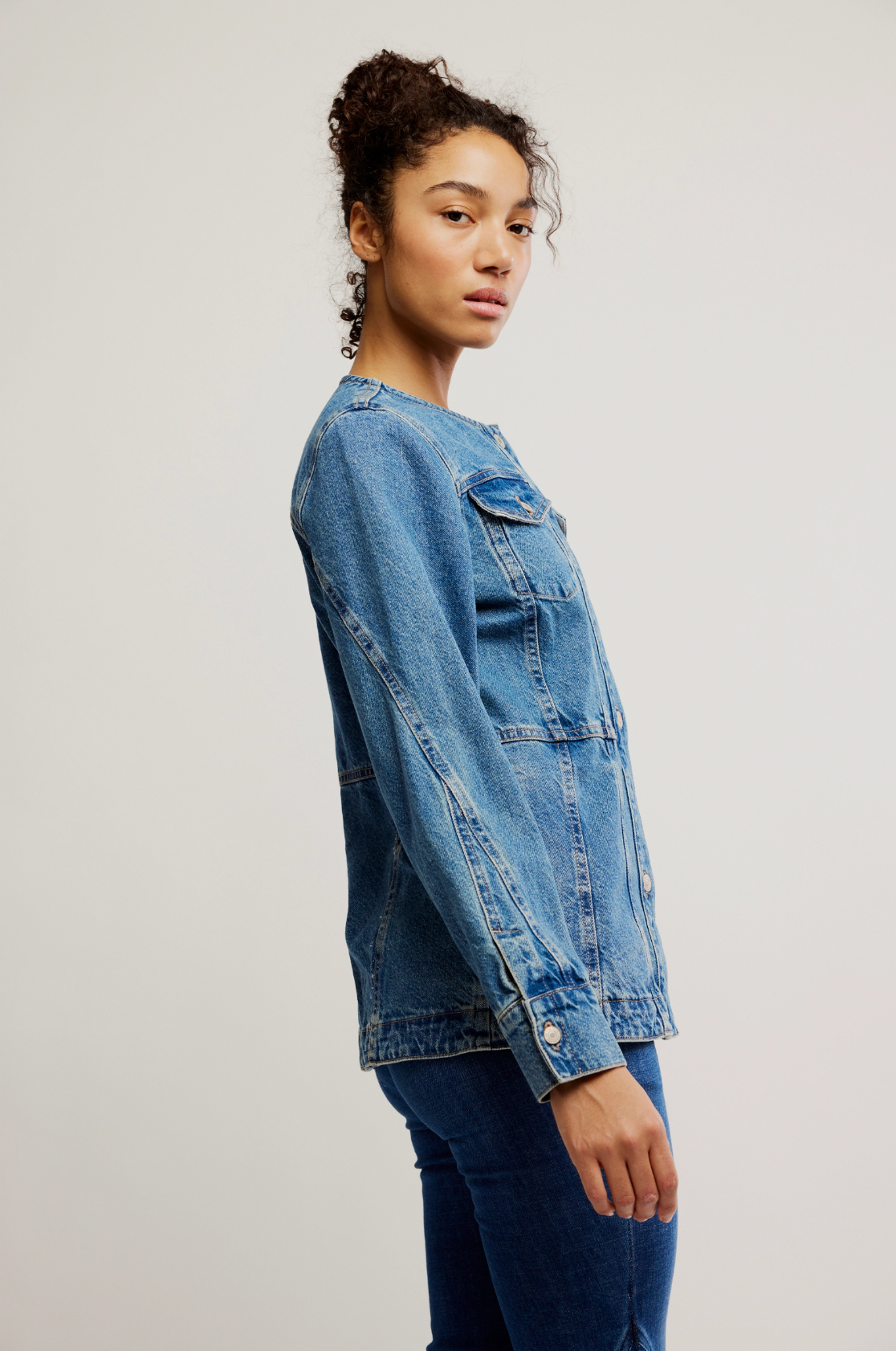 A woman with curly hair models the Free People Lula Denim Jacket with jeans, standing sideways and looking at the camera against a plain light background.