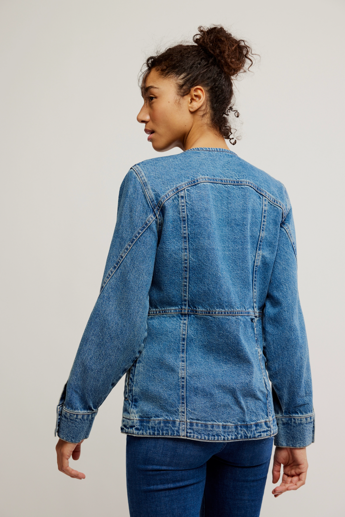 A person with curly hair in a bun is seen from behind wearing the Lula Denim Jacket by Free People and jeans, standing against a plain, light-colored background.