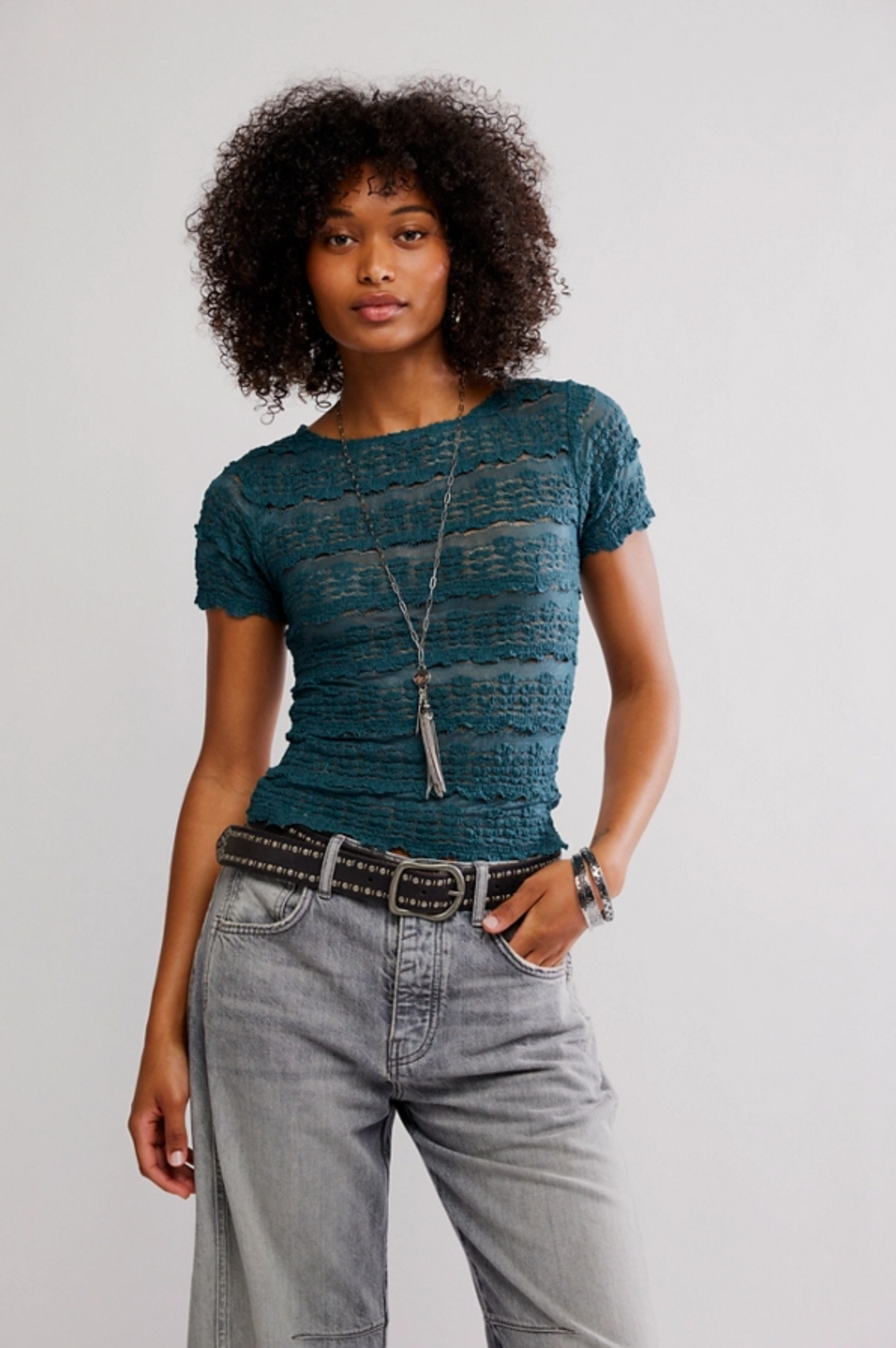 A woman with curly hair poses against a plain background, wearing the Free People Angelina Lace Tee in teal, light gray high-waisted jeans, a black belt, layered necklaces, and bracelets with one hand in her pocket.