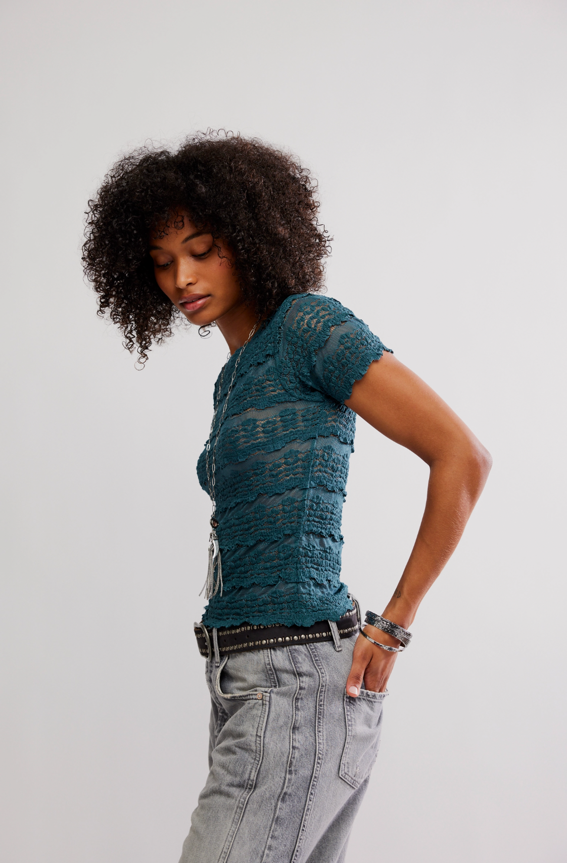 A person with curly hair models the Free People Angelina Lace Tee in teal, featuring a shrunken fit, paired with light gray jeans. They stand sideways with hands in pockets, looking down against a plain light background.