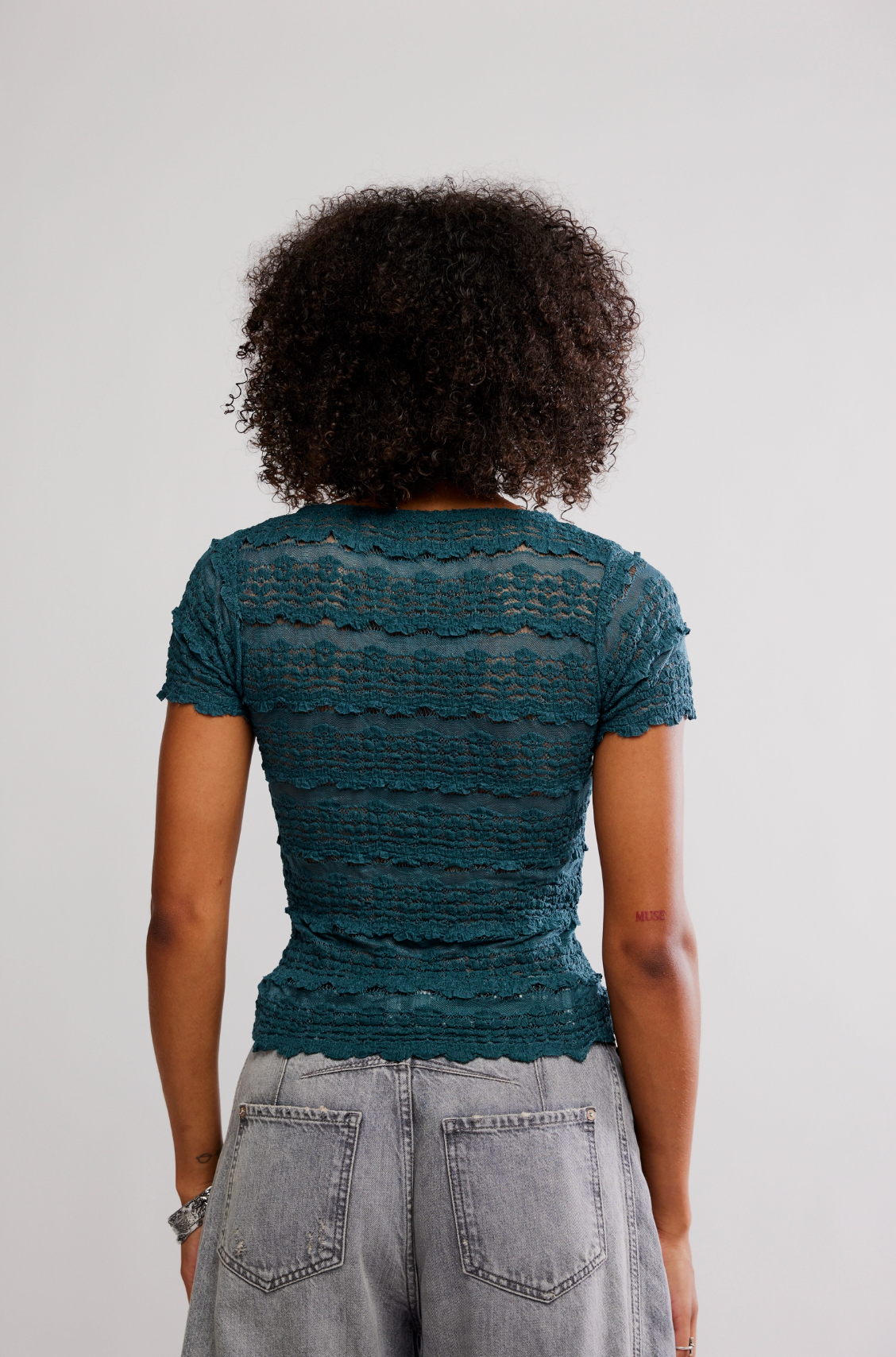 Someone with curly hair is seen from the back wearing the Free People Angelina Lace Tee in teal, featuring a shrunken fit, paired with light gray jeans against a plain background.