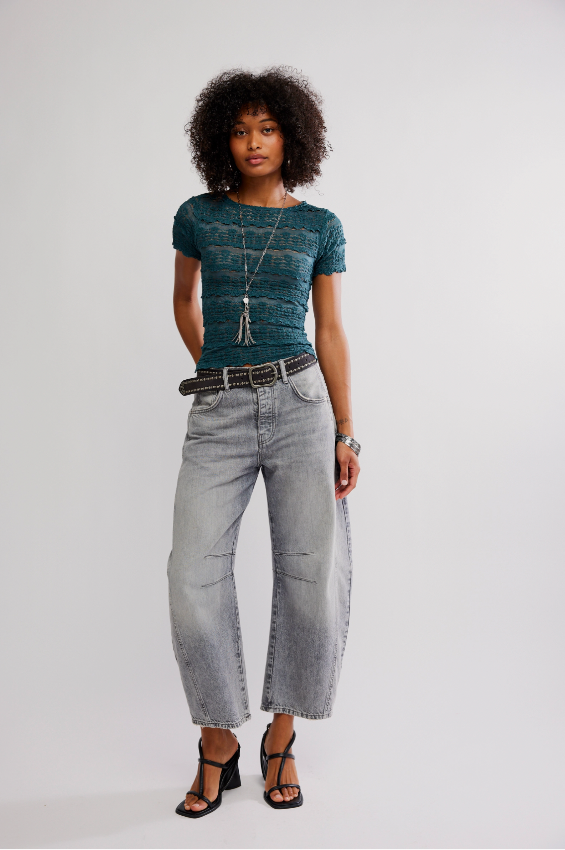 A woman with curly hair stands against a plain light background wearing the Free People Angelina Lace Tee in teal, light gray high-waisted jeans, a black belt, long necklace, bracelets, and black heeled sandals.