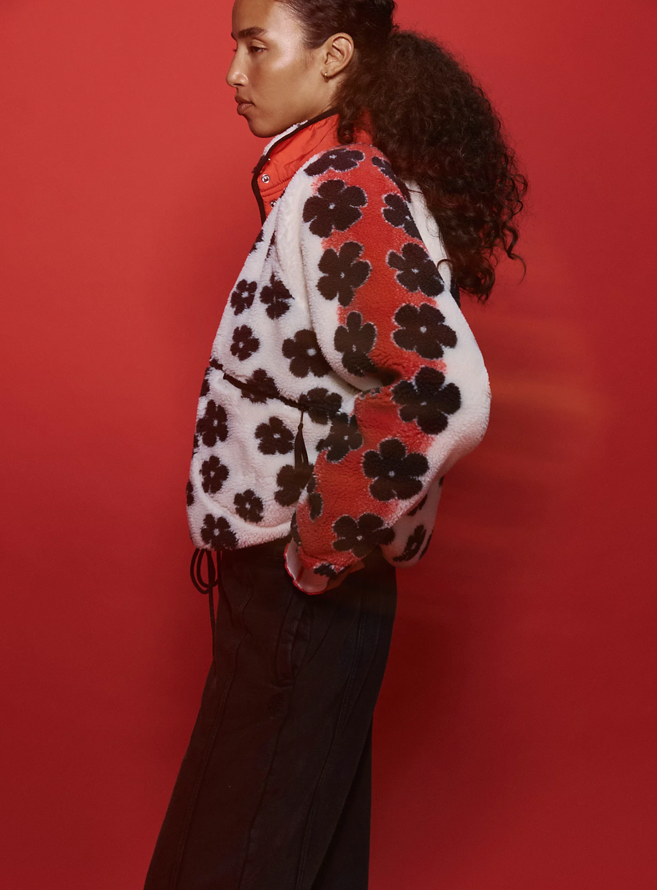 A person with curly hair stands in profile against a red background, wearing Free People's Hit The Slopes Printed Fleece Jacket featuring black floral patterns on white and red panels, styled with dark pants.