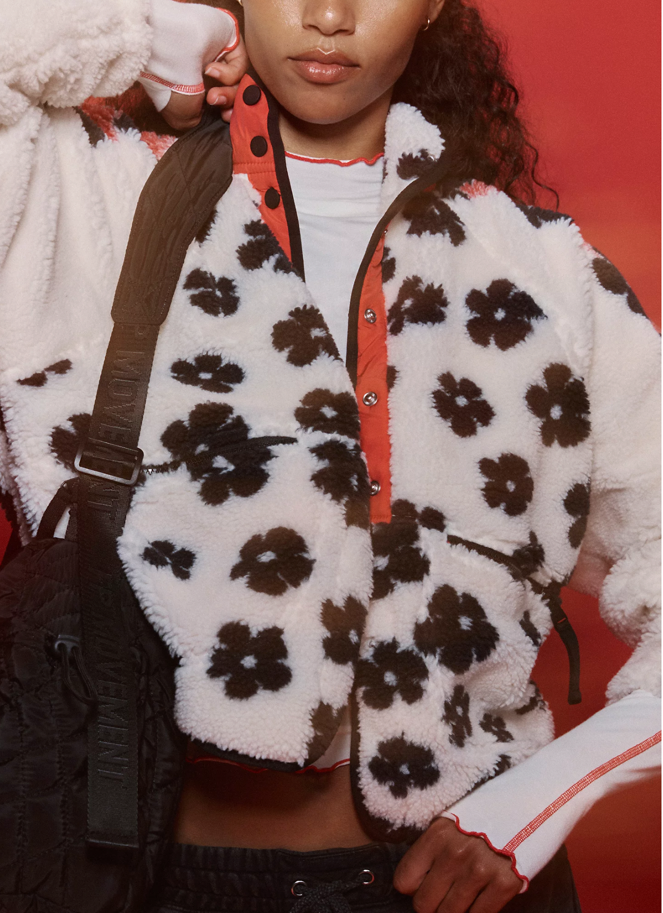 A woman wearing the Free People Hit The Slopes Printed Fleece Jacket, a white fleece with black flower patterns, poses against a red background with an orange shirt and black shoulder bag, her right arm raised and partly covering her face.