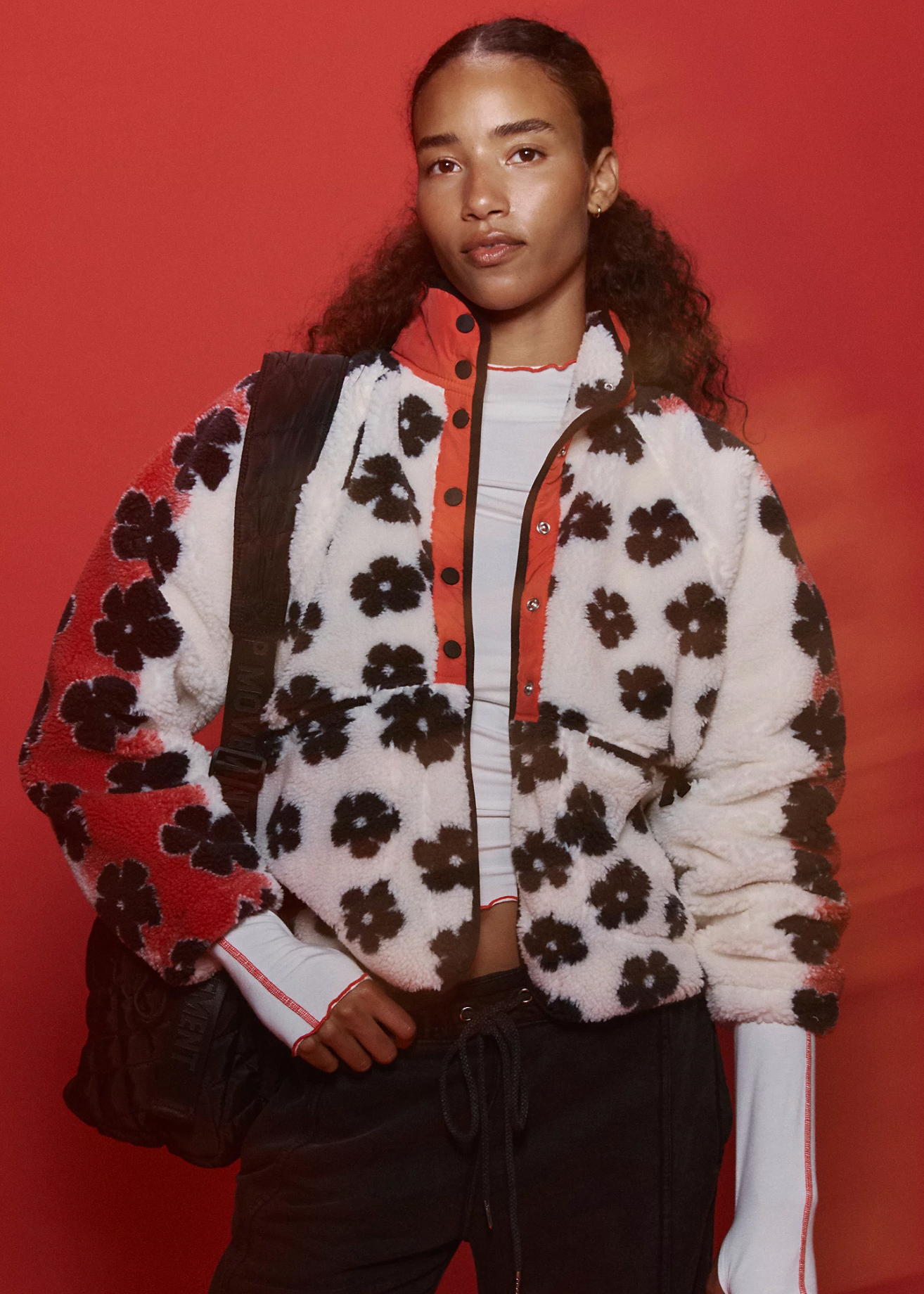 A person stands against a red background, modeling streetwear with Free People's Hit The Slopes Printed Fleece Jacket—white and red with black flower patterns—layered over a light shirt, paired with black pants and a black shoulder bag.