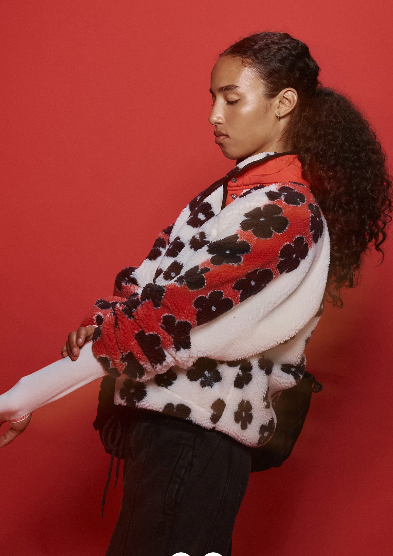 A person with curly hair in a ponytail wears the Free People Hit The Slopes Printed Fleece Jacket, featuring black and red floral prints, standing against a solid red background and looking downward.