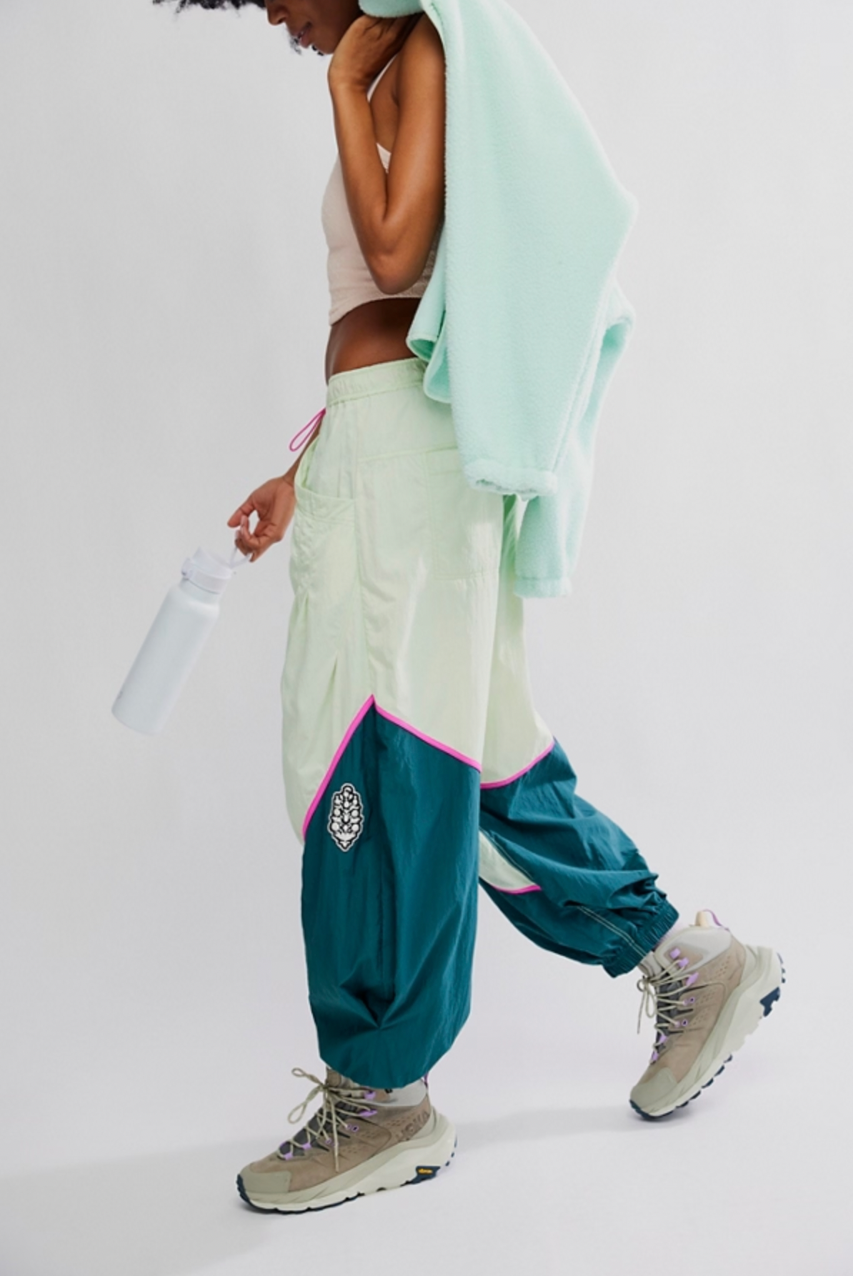 Wearing Free People’s Free Flow Pant in Spring Meadow, a person pairs the colorblock track pants with a beige tank top, beige sneakers, and drapes a light green fleece over their shoulder while holding a water bottle against a plain background.