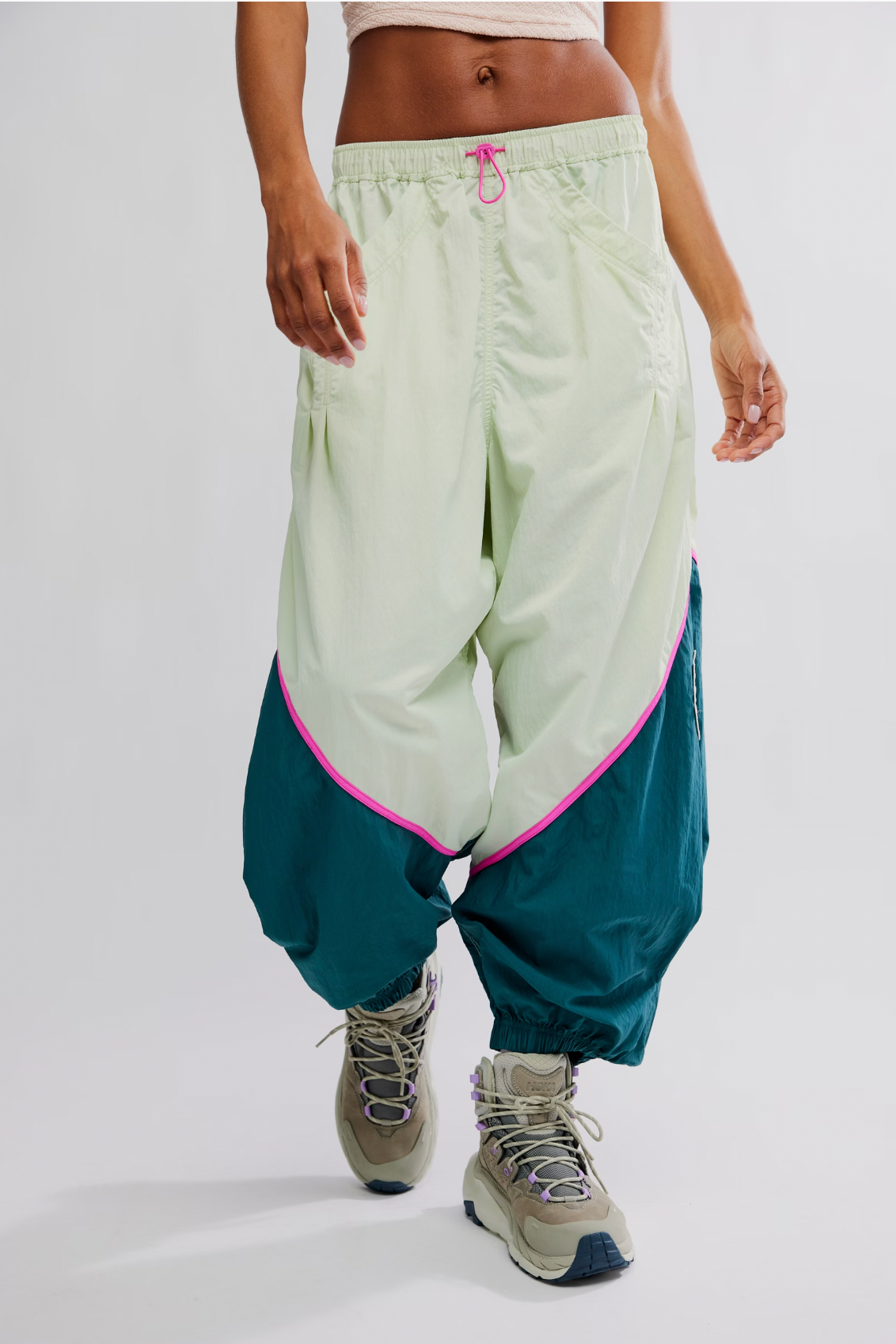 Someone wearing the Free People Free Flow Pant in Spring Meadow—a slouchy, ankle-length nylon pant with a green and teal colorblock and pink trim—paired with chunky gray sneakers, walks forward against a plain light background.