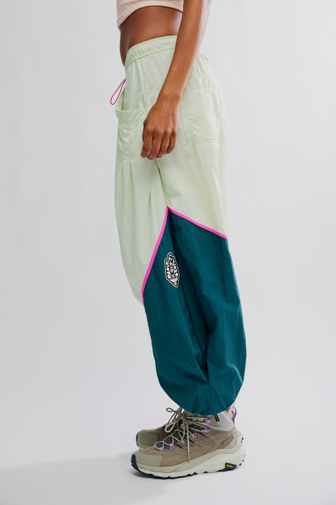 A person in Free People’s Free Flow Pant in Spring Meadow, featuring a relaxed fit and colorblock design in light green and teal with pink trim, pairs the look with chunky beige sneakers and a light beige crop top against a white background.