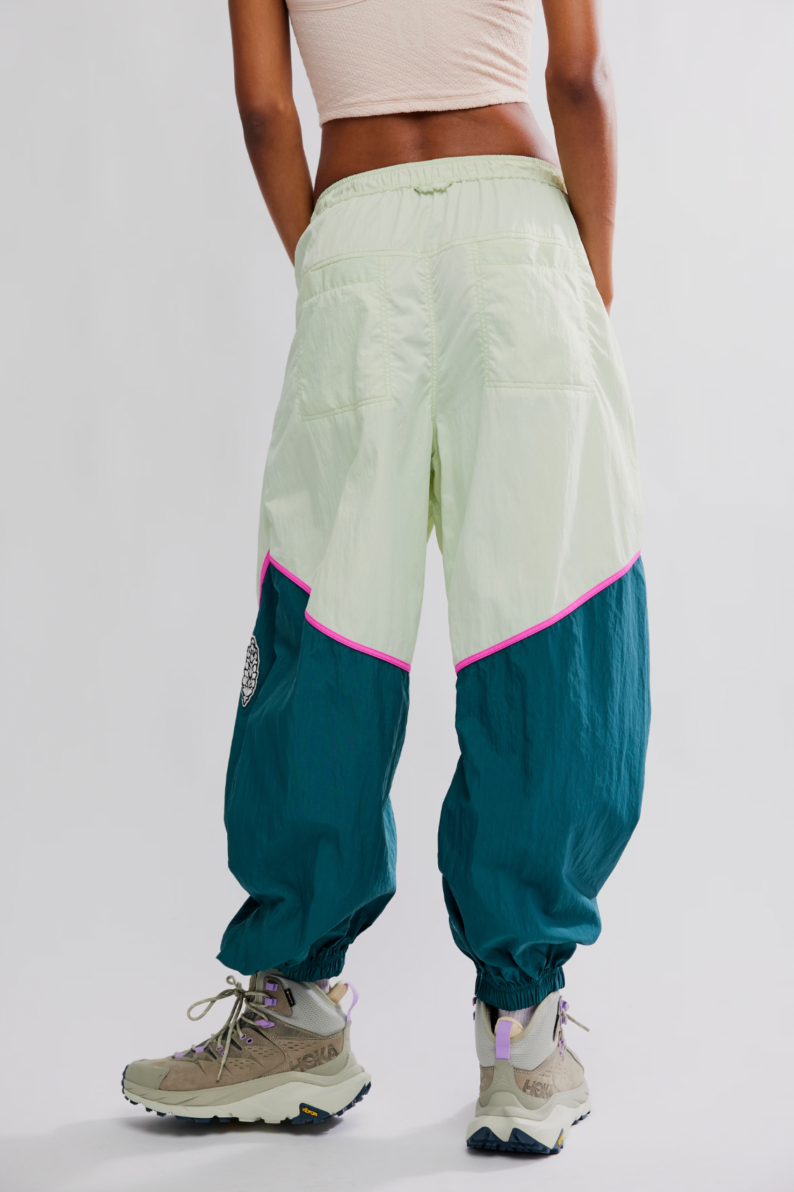A person stands facing away against a plain light background, wearing Free People’s Free Flow Pant in Spring Meadow—sage green and teal colorblock track pants with a pink stripe—paired with a beige crop top and trail sneakers.