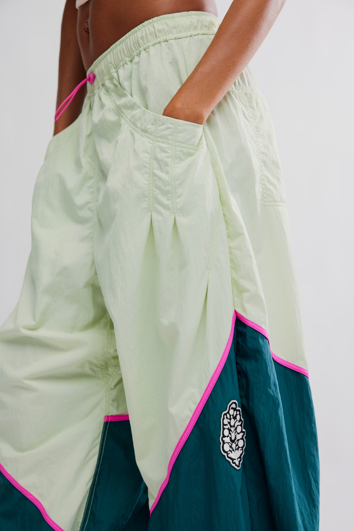 A person wearing Free People's Free Flow Pant in Spring Meadow—a slouchy nylon colorblock style in light green and dark teal with pink trim and a decorative patch—stands with one hand in their pocket.