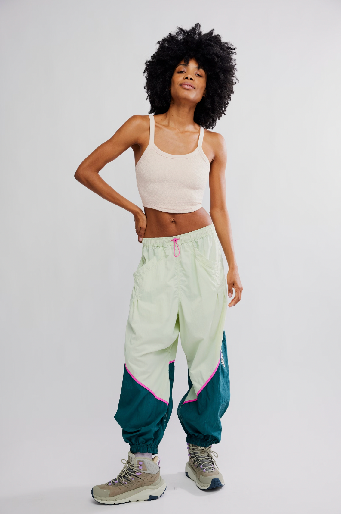 A woman with curly hair and a bright smile wears the Free Flow Pant in Spring Meadow by Free People, styled with a light crop top and hiking boots, standing against a plain light background.