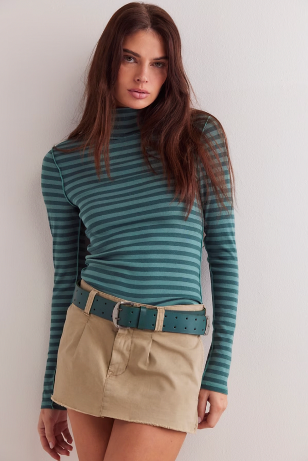 A woman with long brown hair wears the Free People Good Good Turtle Stripe Top in Rainforest, paired with a tan mini skirt and wide teal belt, standing against a white wall.