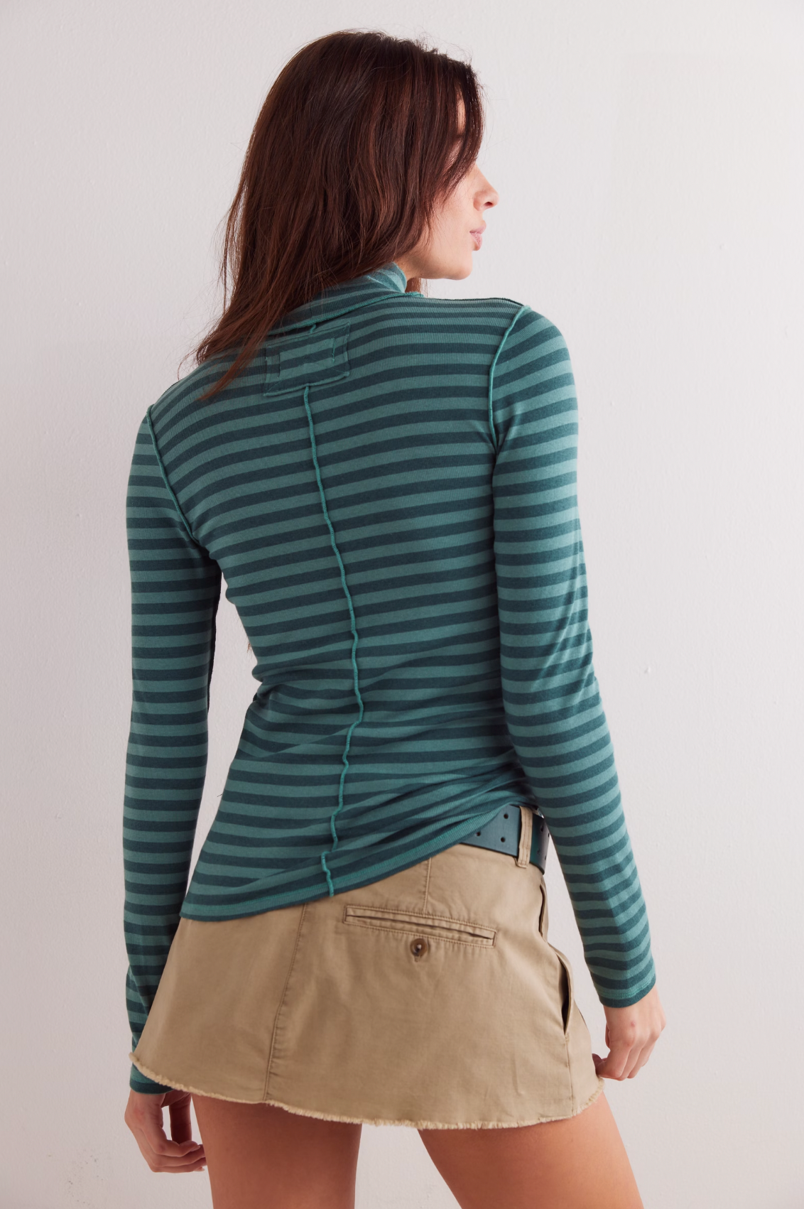 A woman with brown hair, seen from the back, wears the Free People Good Good Turtle Stripe Top in Rainforest and a beige mini skirt against a plain white wall.