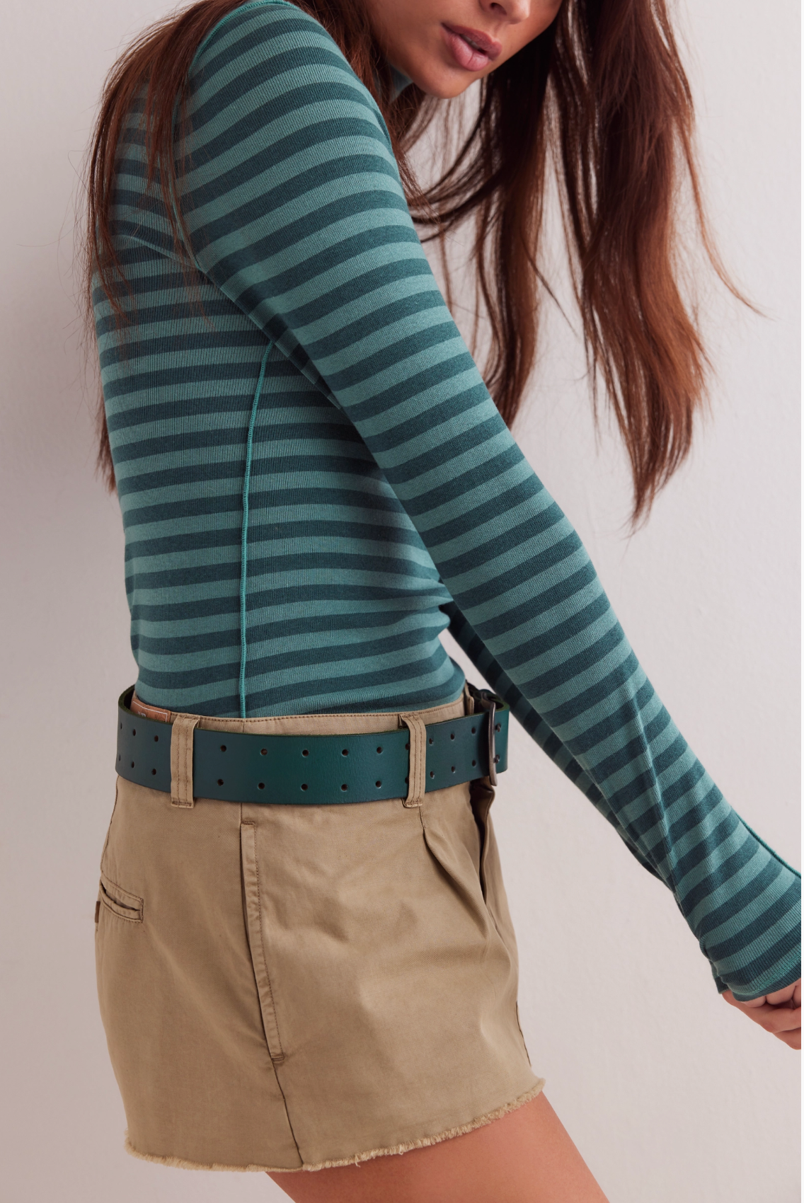 A woman with long brown hair wears the Good Good Turtle Stripe Top in Rainforest by Free People, tan frayed-hem shorts, and a wide green belt. She stands against a plain light background.
