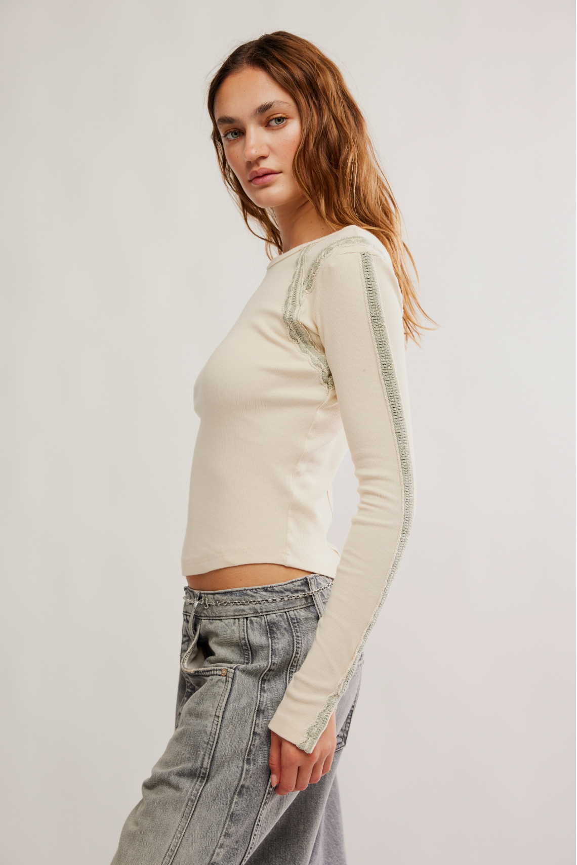 A woman stands sideways in the Caravan Lost Love Long-Sleeve Top, featuring a relaxed fit, off-white ribbed fabric, lace-trimmed sleeves, and light-wash jeans, set against a plain light background.