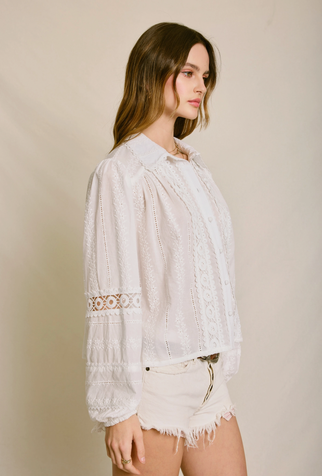 A woman stands in profile against a neutral background, wearing the Dress Forum Sunbeam Eyelet Blouse and white frayed shorts. Her hair is loose and she has a calm expression.