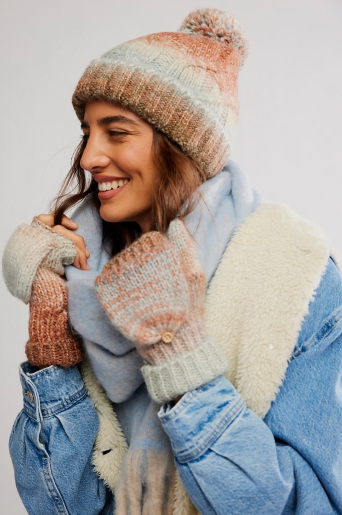 A smiling woman in the Free People Rainbow Run Hat and Glove Set in Blue Combo, featuring convertible mittens, a light blue scarf, a sherpa-lined jacket, and denim outerwear, faces left against a plain background.