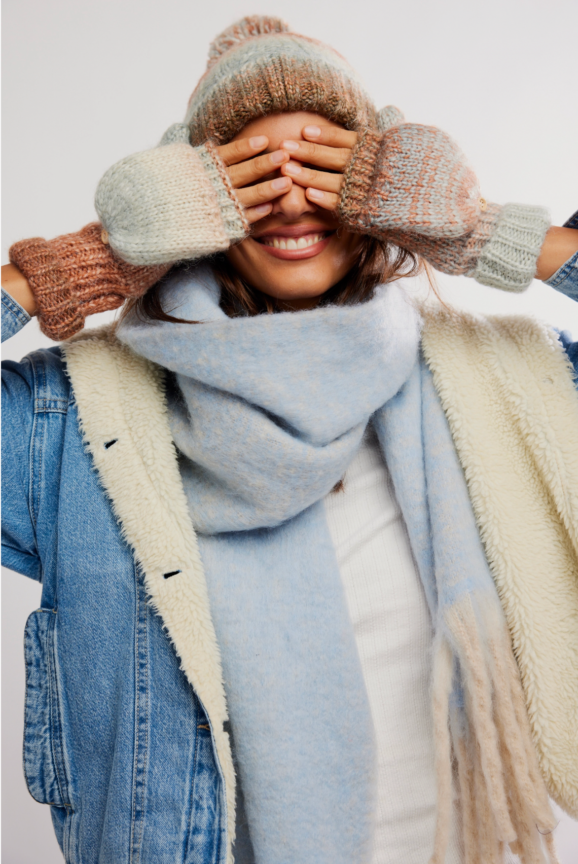 A person wearing the Free People Rainbow Run Hat and Glove Set in Blue Combo, featuring multicolored convertible mittens, a blue scarf, and a shearling-lined denim jacket, smiles while playfully covering their eyes with their hands.