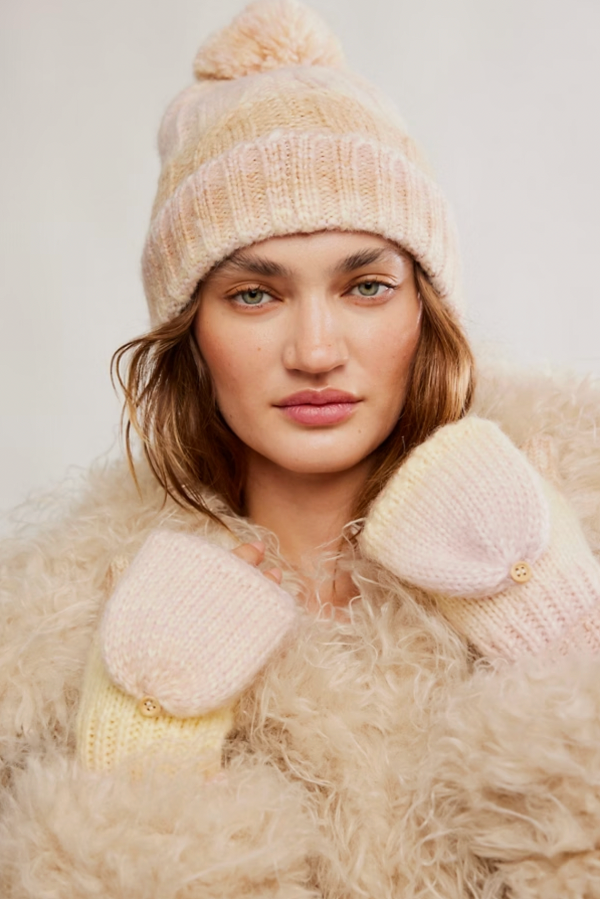 A woman wearing the Free People Rainbow Run Hat and Glove Set in Ivory, paired with a fluffy cream-colored coat, looks at the camera against a plain background.