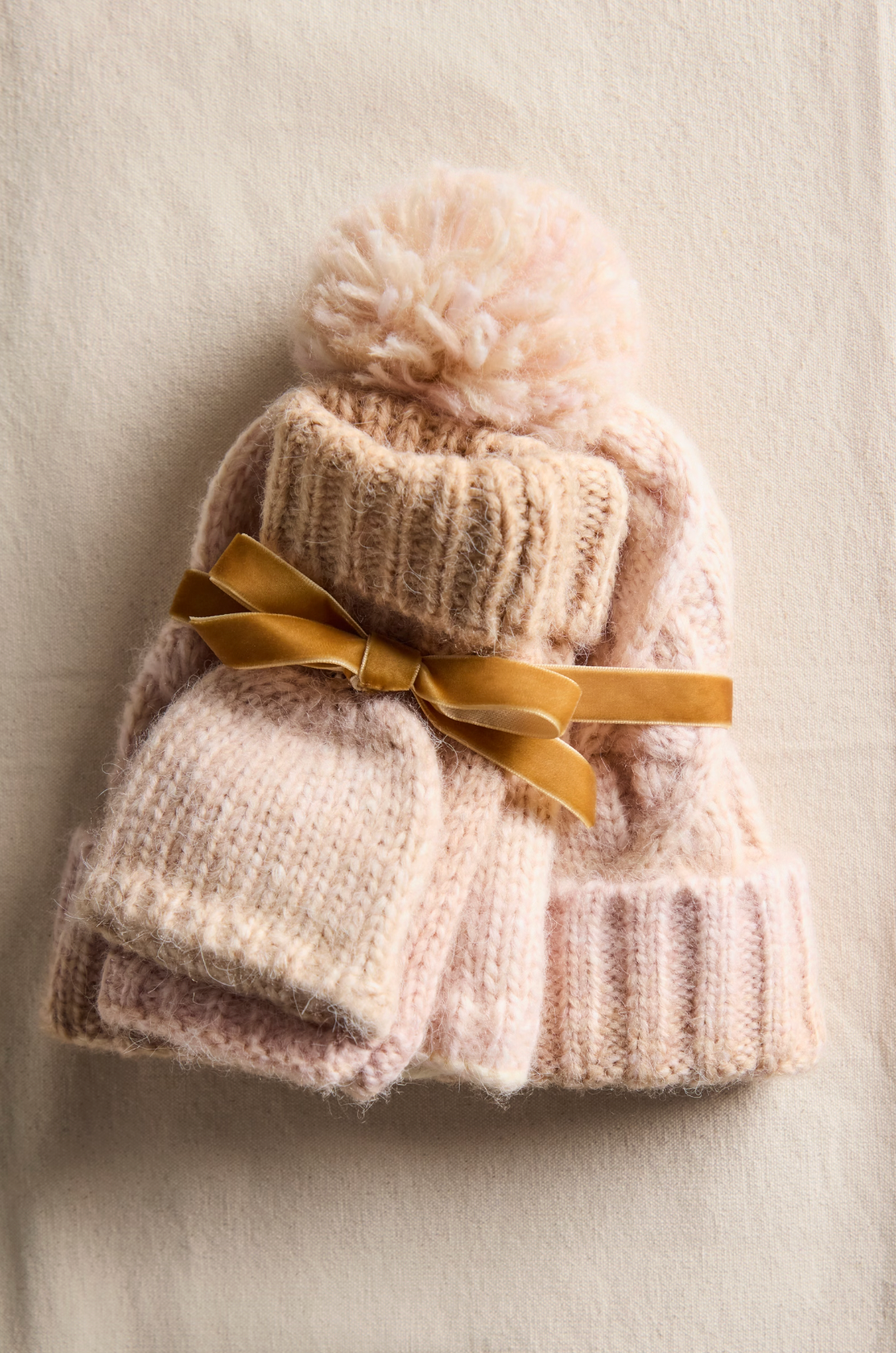 The Free People Rainbow Run Hat and Glove Set in Ivory includes a knit hat, scarf, and convertible mittens, stacked and tied with a gold velvet ribbon, shown on a cream fabric background.