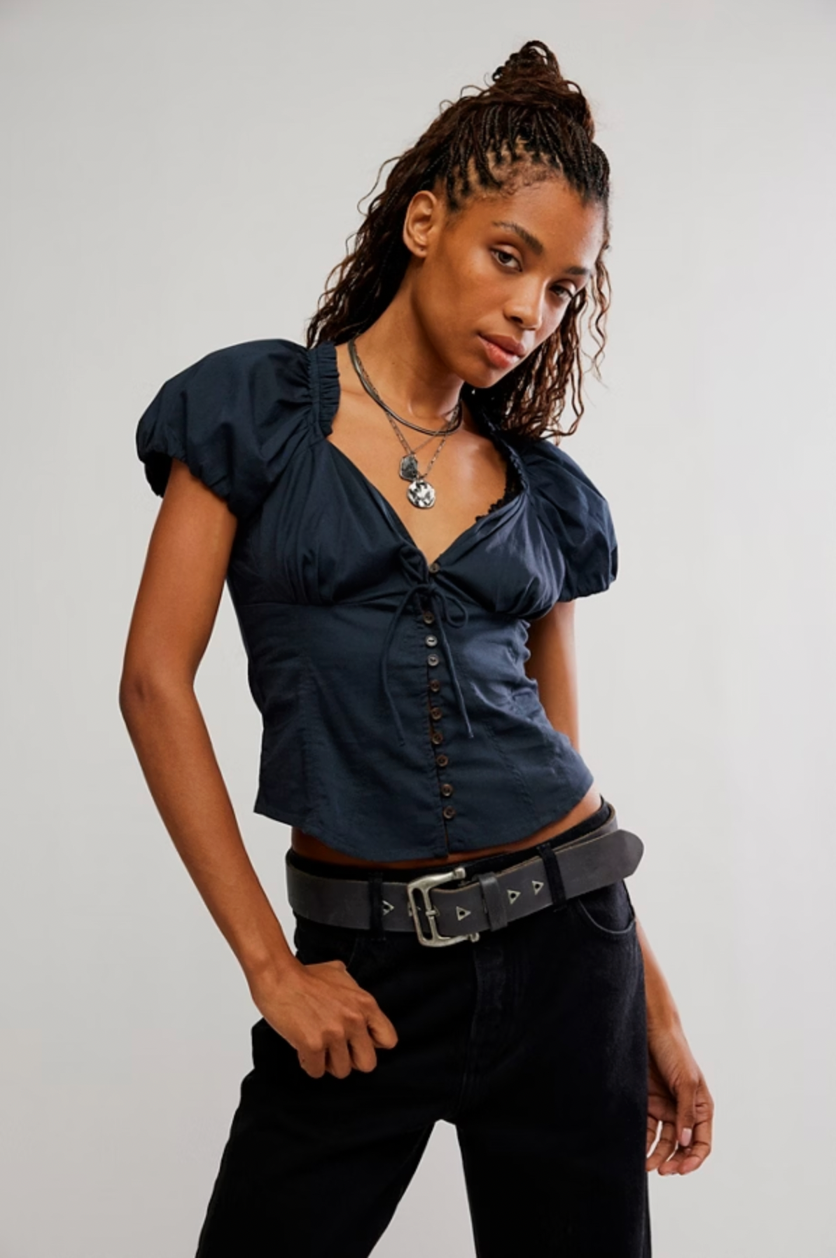 A woman with braided hair models Caravan's The Staple Blouse in navy with puff sleeves and ruffled trim, paired with black pants, a wide black belt, and layered necklaces against a plain light background.