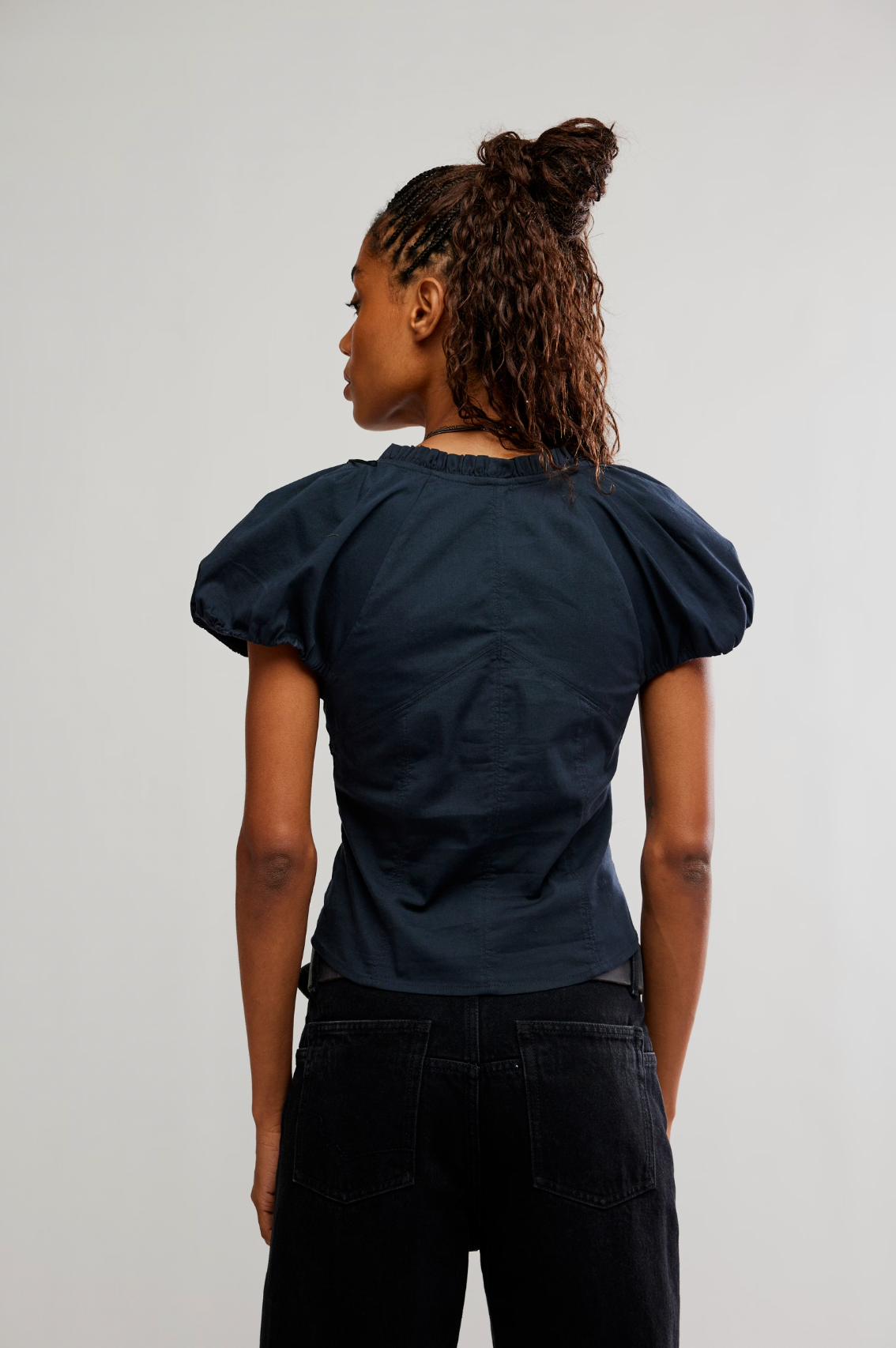 A woman with loose curls in a half-up bun wears Caravan's The Staple Blouse in navy with puff sleeves and black jeans, standing against a plain light background.