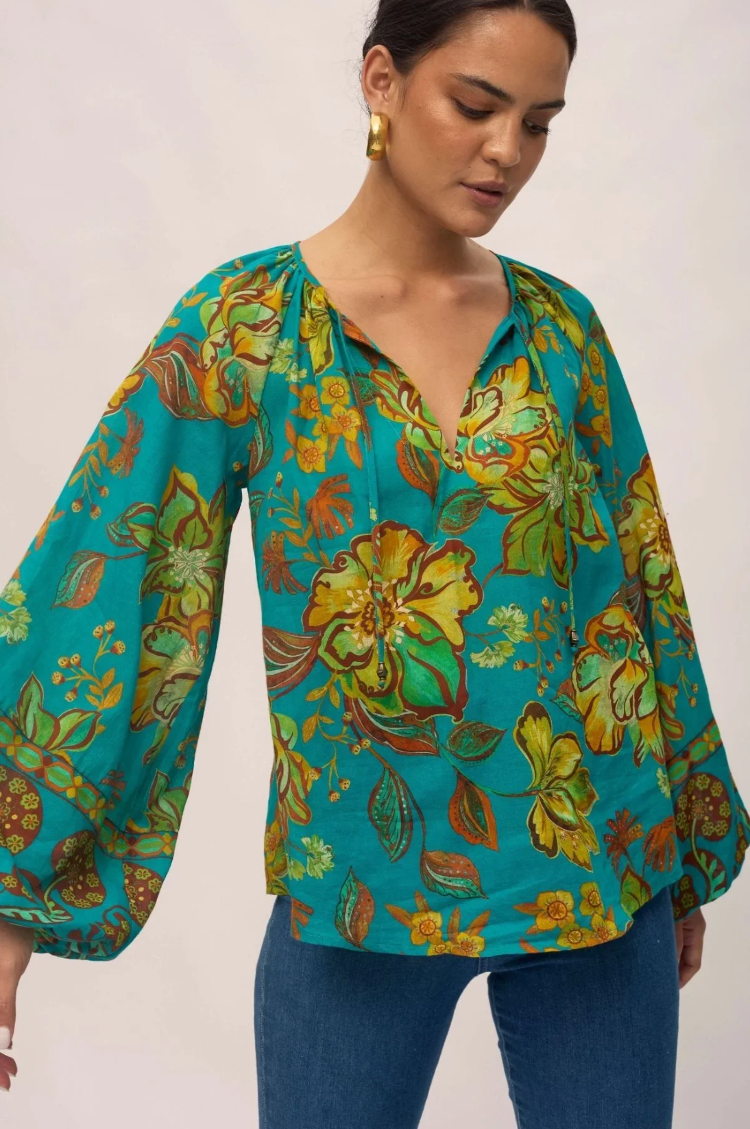 A woman wears the Nine Lives Bazaar Flora Blouse - Peacock, featuring a bold yellow and green floral pattern, raglan poet sleeves, and a V-neckline. She pairs it with blue jeans and gold earrings against a plain light background.
