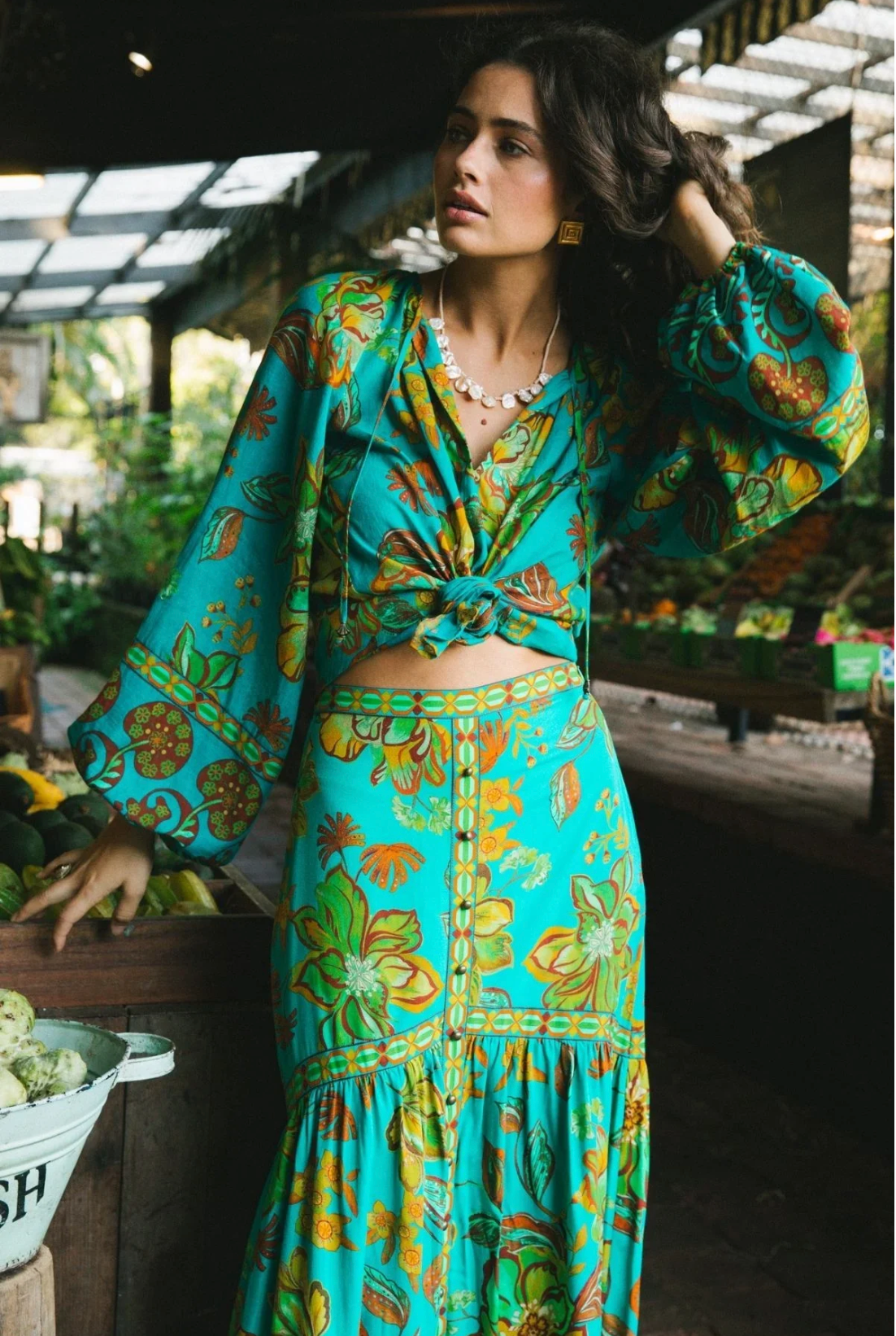A woman stands in a market, wearing a bright turquoise skirt and the Nine Lives Bazaar Flora Blouse - Peacock, featuring an intricate floral and fruit pattern. Her wavy hair and the blouse’s raglan poet sleeves highlight her bohemian charm.