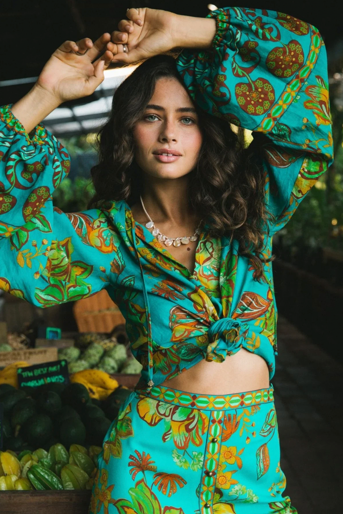 A woman with wavy brown hair confidently models the Nine Lives Bazaar Flora Blouse - Peacock, featuring raglan poet sleeves, while standing before a market stall filled with fresh produce.
