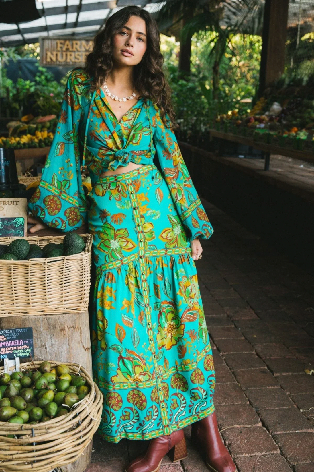 Wearing the Nine Lives Bazaar Flora Blouse in Peacock, a woman with vibrant raglan poet sleeves and red boots stands by baskets of fruit at an outdoor market, surrounded by fresh produce and greenery.
