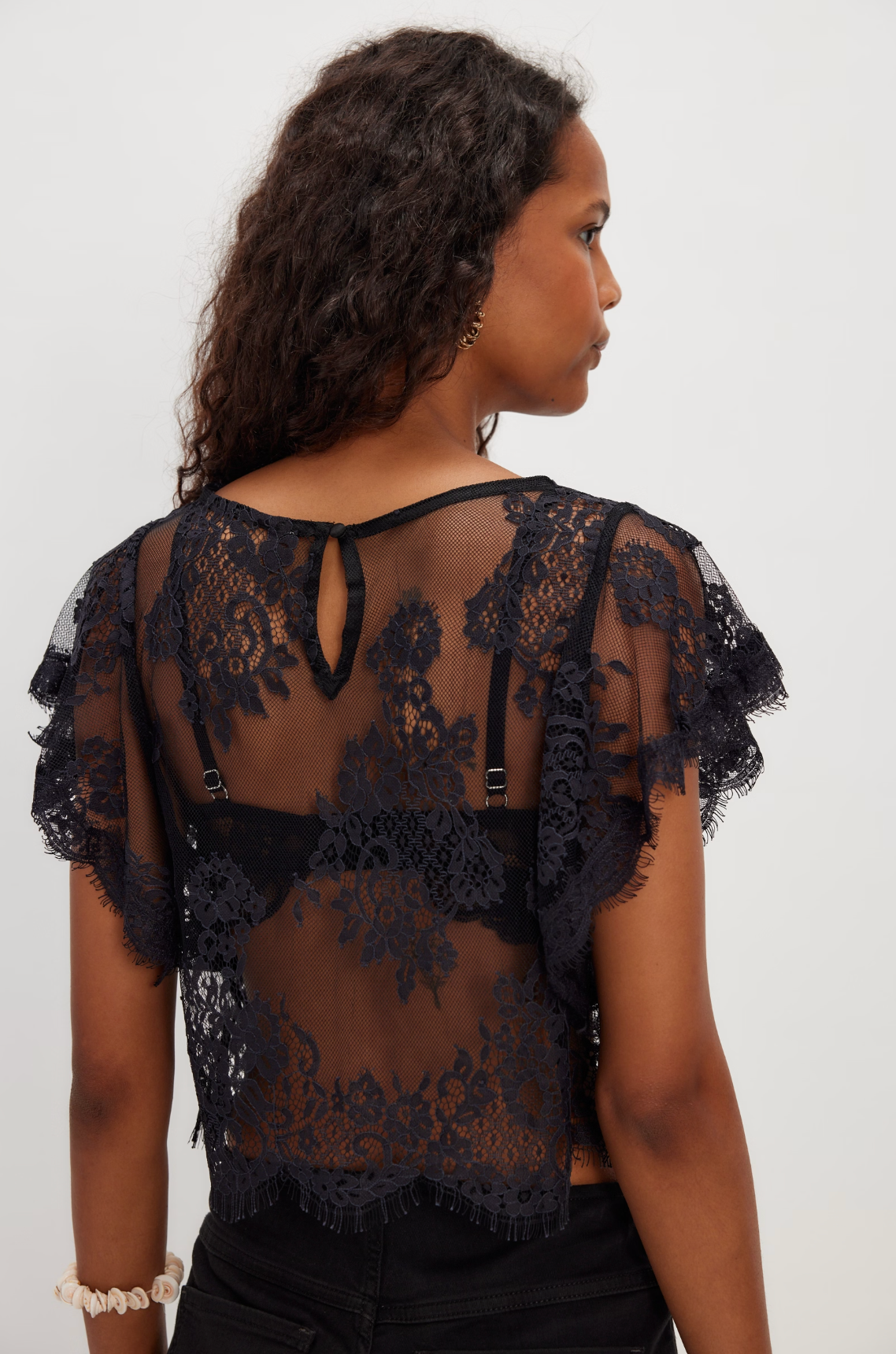 Woman wearing a black Lunar Ash Lace Top featuring floral lace and sheer fabric from the back view.