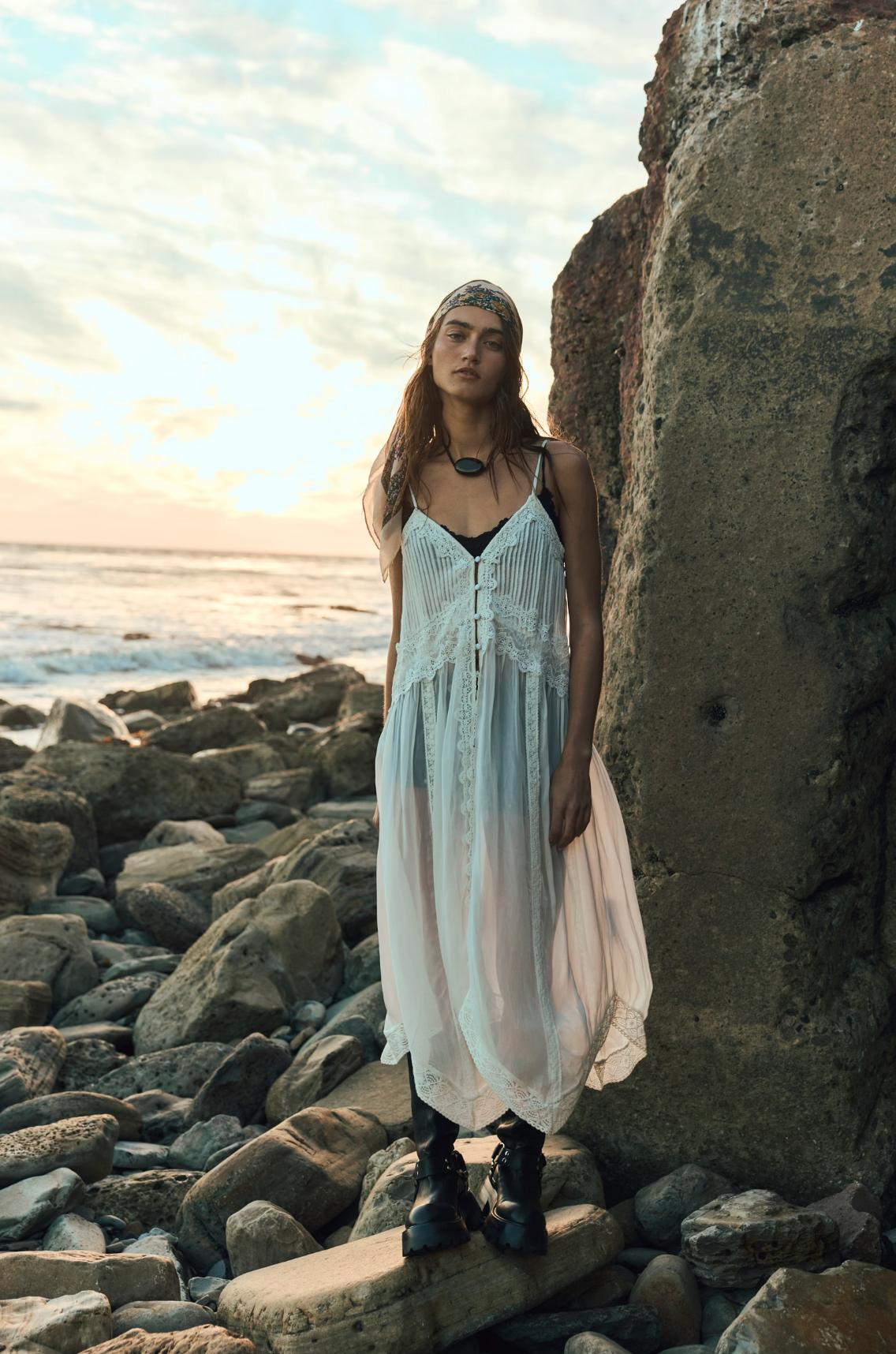 Woman wearing a flowing Snowbird Maxi Top with lace details standing on rocky shore at sunset.