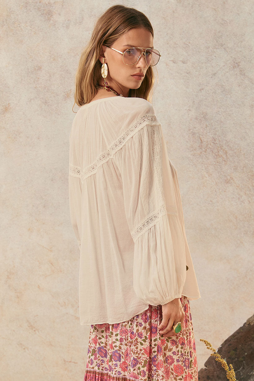 A woman with long hair and oversized glasses wears the Teodora Blouse - White Smoke by Spell, crafted from LENZING ECOVERO Viscose with lace details. She pairs it with statement earrings and a floral skirt, glancing over her shoulder.