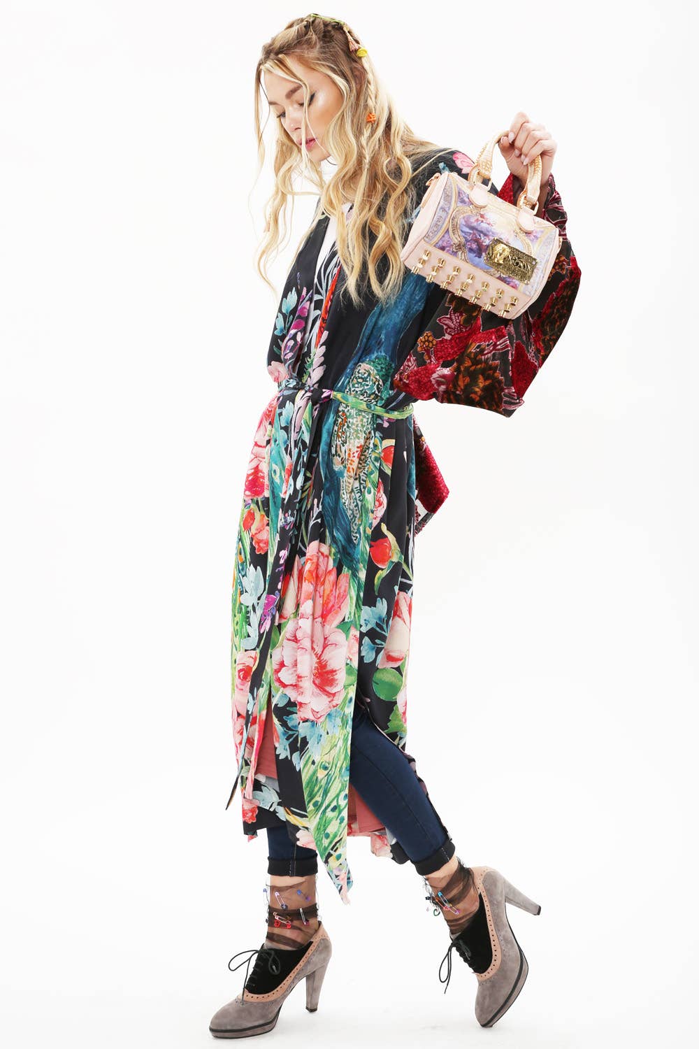 A woman stands against a white background wearing the Aratta Sweet Fantasy Kimono with burnout velvet sleeves over dark pants. She holds a small pink handbag, wears gray heels, and styles her long blonde hair with clips.
