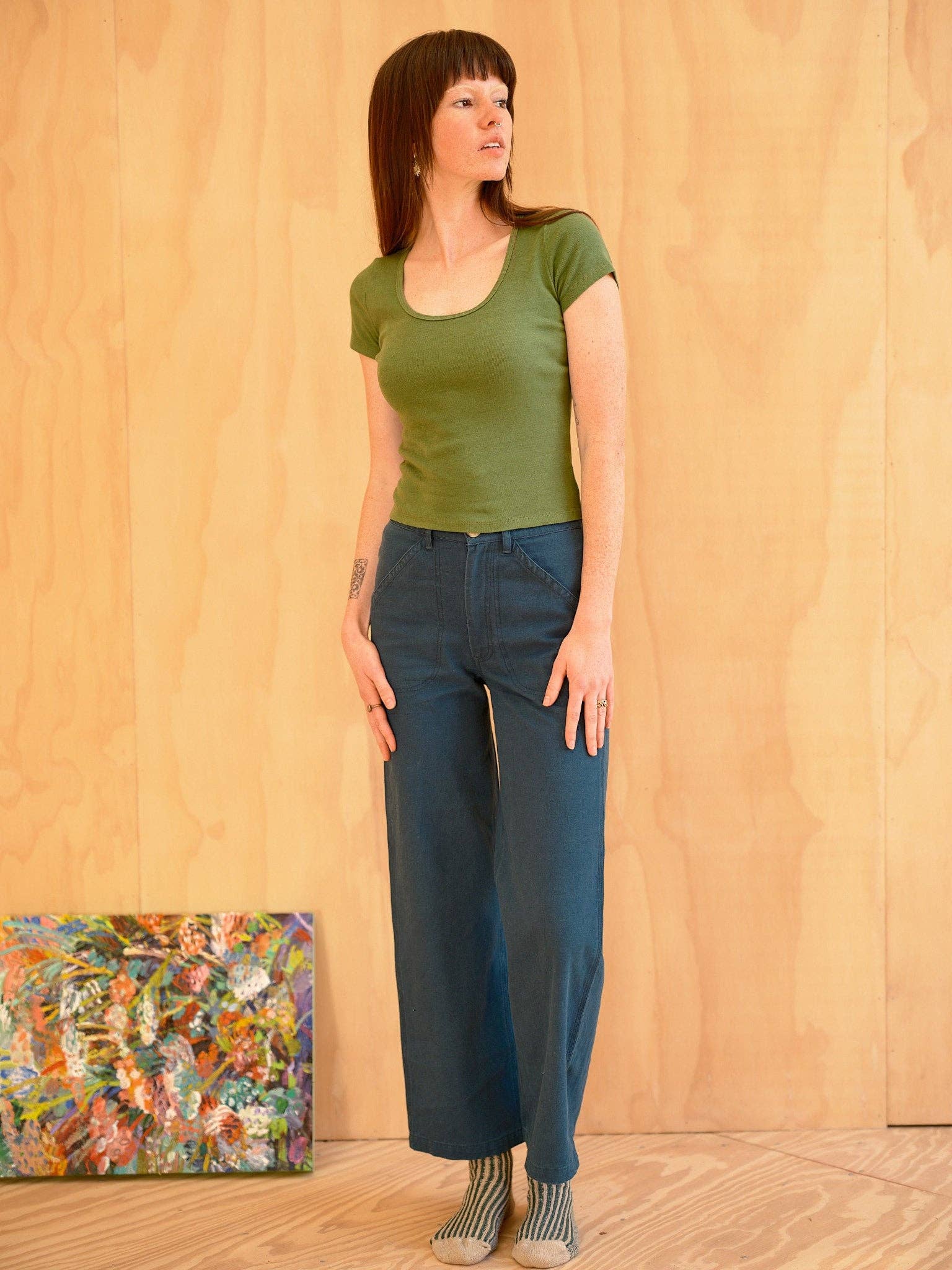 A woman with straight brown hair wears the Mollusk Scoop Ribsy Tee in green and blue wide-leg pants, standing indoors on a wooden floor near a colorful abstract painting.