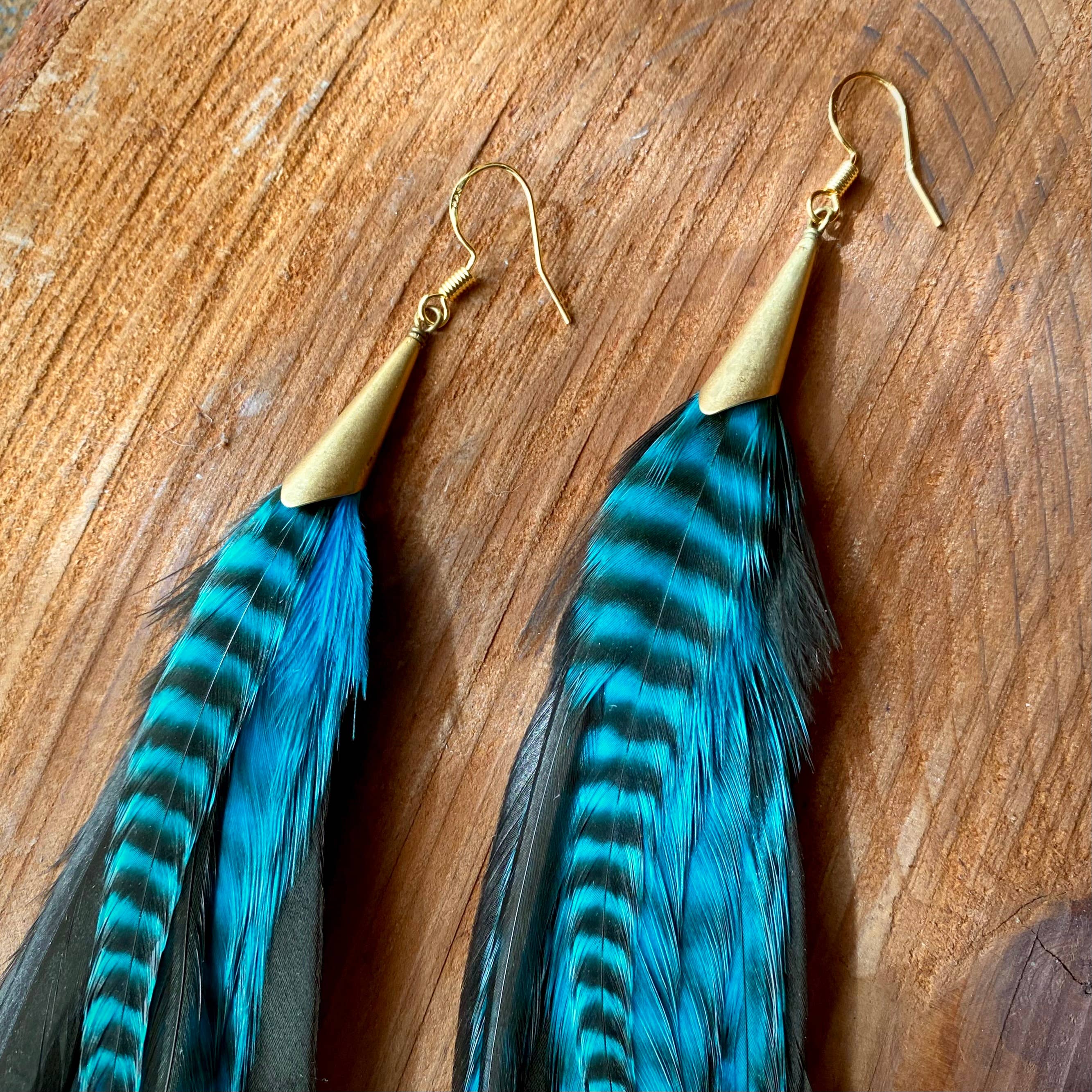 ASTALI Long Feather & Cone Earrings in Turquoise Grizzly & Black/Gold feature gold hooks and caps with sustainably sourced striped feathers, shown on a wooden surface.