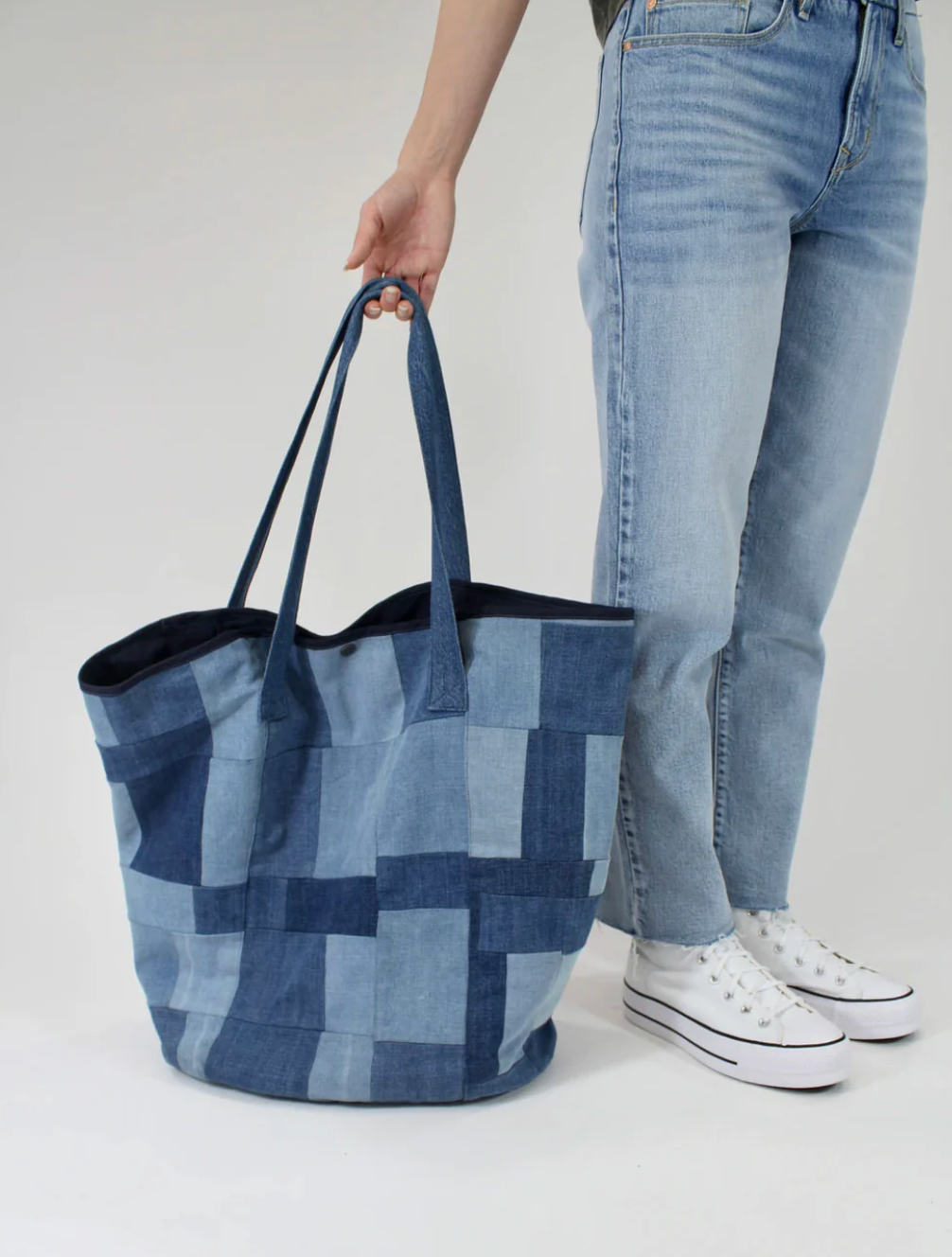 A person in light blue jeans and white sneakers holds the PATCHWORK TOTE by Porter Blue Apparel, crafted from denim in various blue shades. The background is plain and light-colored.