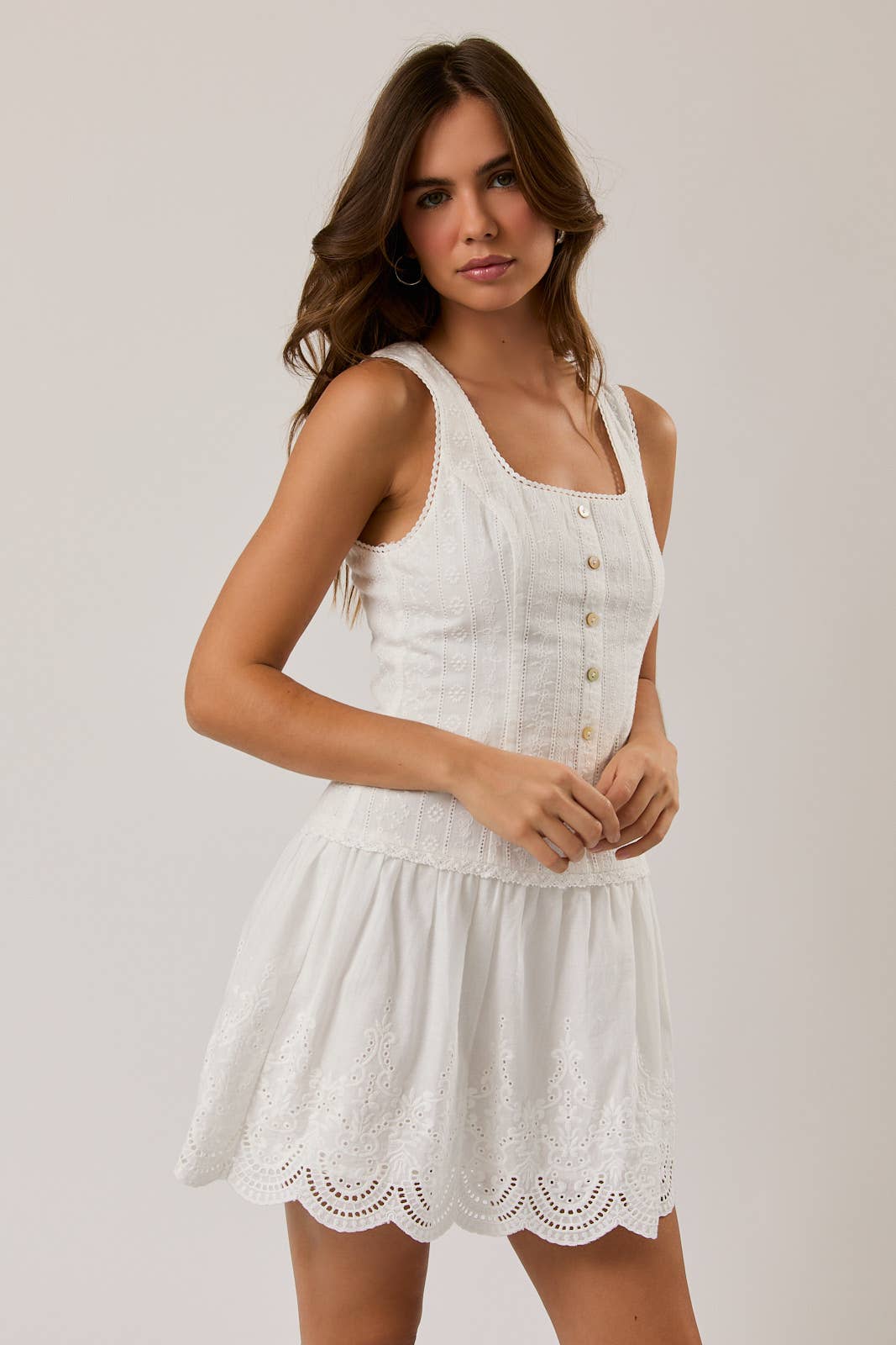 Young woman wearing a white sleeveless eyelet mini dress with button front and scalloped hem detail