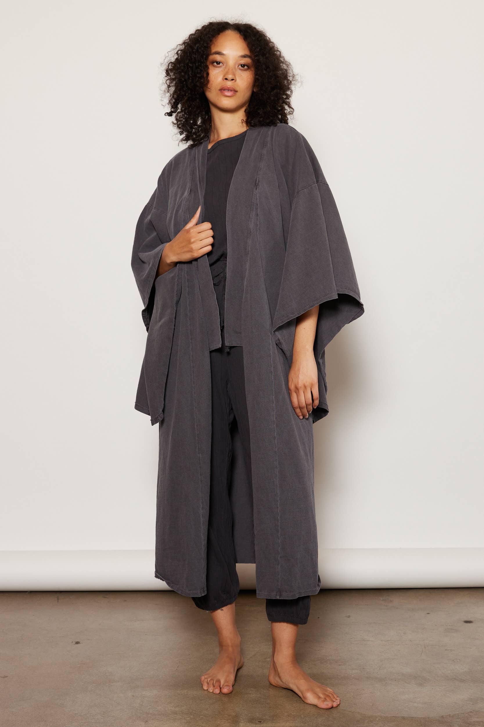 A woman with curly hair stands barefoot on a concrete floor, wearing the Dawn Robe by People Of Leisure—an ethically made, long dark gray organic cotton kimono—over a black outfit, posed against a plain off-white background.