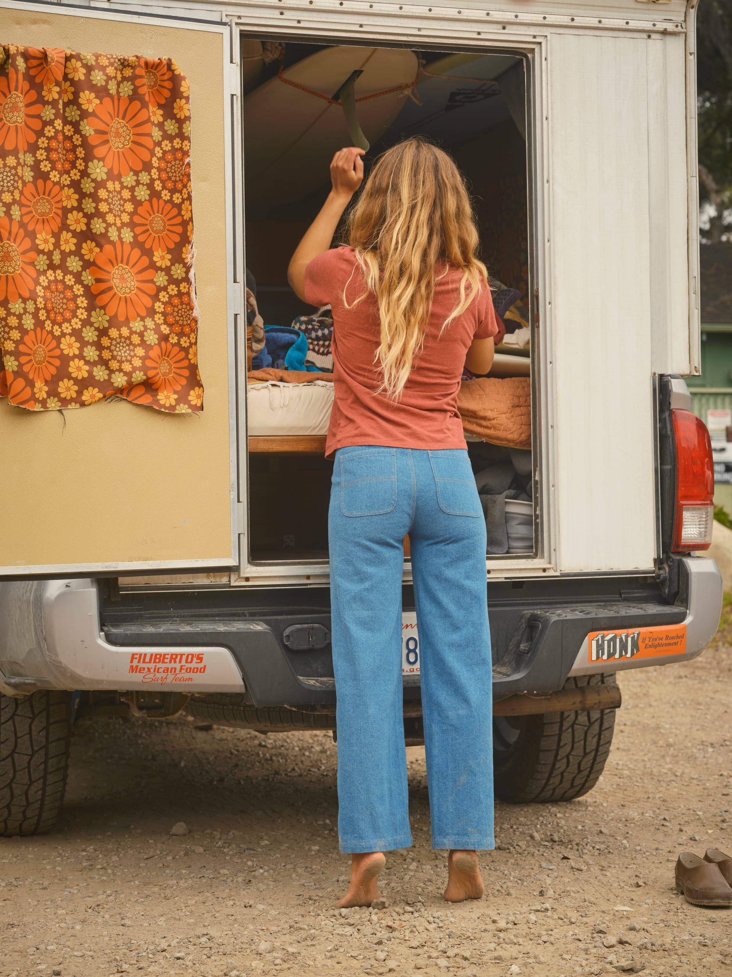 A barefoot woman with long, wavy hair wearing a red shirt and Mollusk's Painter Pants in Indigo stands at the back of a camper van with floral curtains, reaching inside while the van is parked on dirt.