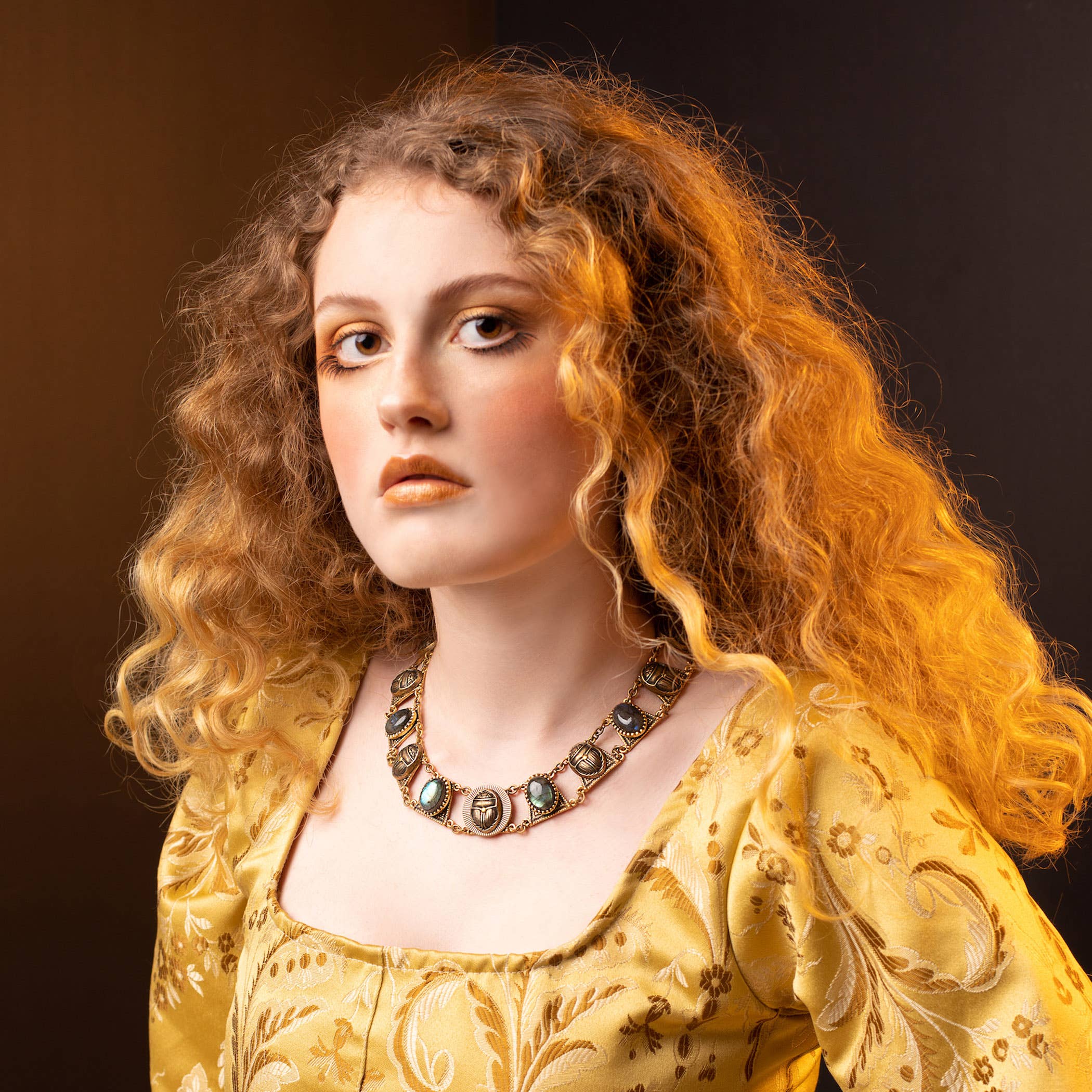 Woman with curly hair wearing a golden dress and Sacred Scarab Collar Necklace featuring turquoise and brass accents.